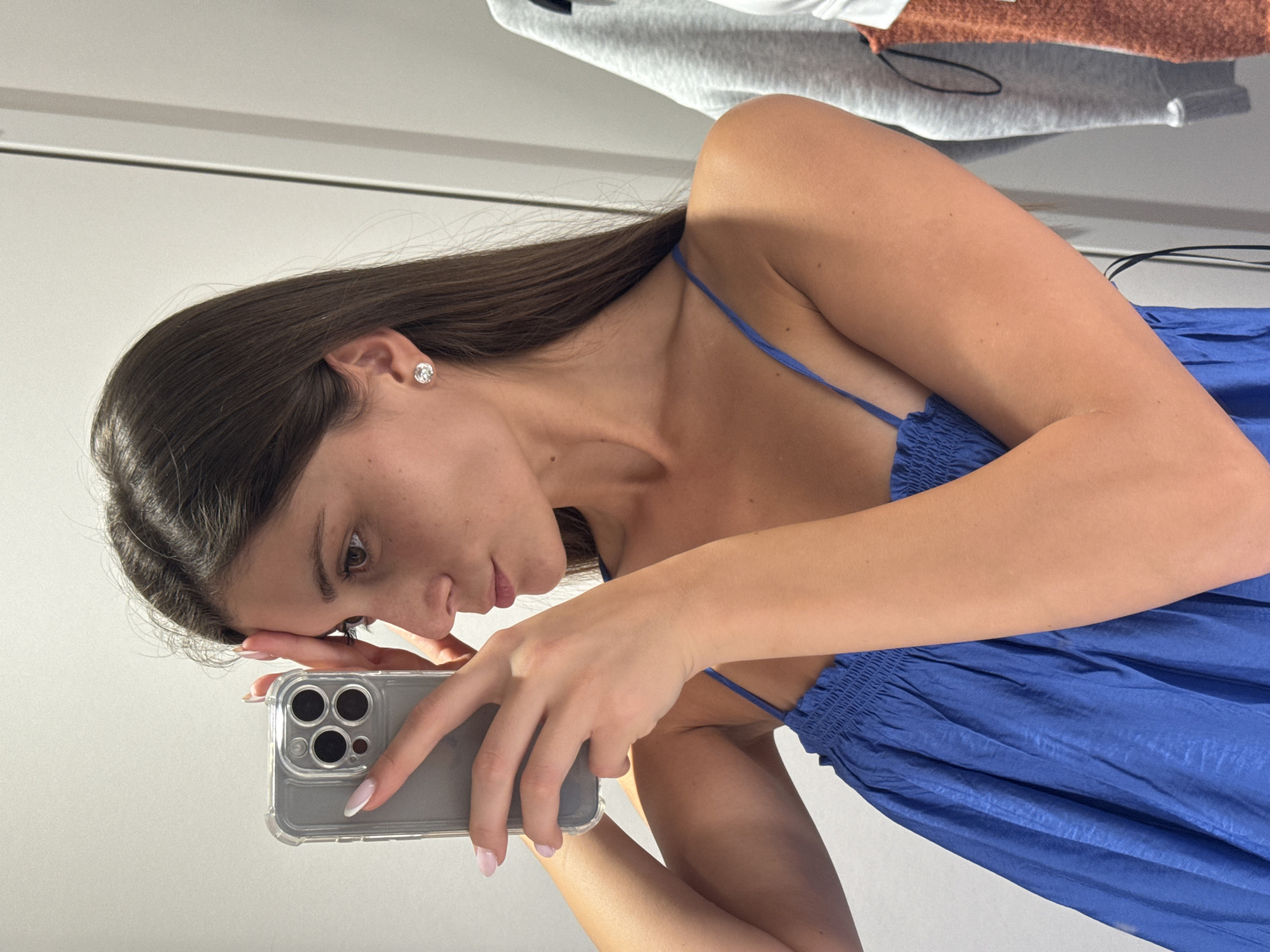 A young woman with long, dark hair takes a mirror selfie in what appears to be a fitting room or changing area. She is wearing a blue dress with spaghetti straps and a smocked bodice. Her left hand is holding a smartphone up to take the picture, and her right hand is resting on her forehead. She is wearing a diamond stud earring in her right ear. The background is a plain, light-colored wall with a doorway or partition visible on the right. A grey sweatshirt and an orange textured garment are hanging on a rack to the right of the woman. The lighting is bright and natural. The woman's facial features are partially obscured by her hand and the phone. There is no information available about the location in Burgas, Bulgaria.