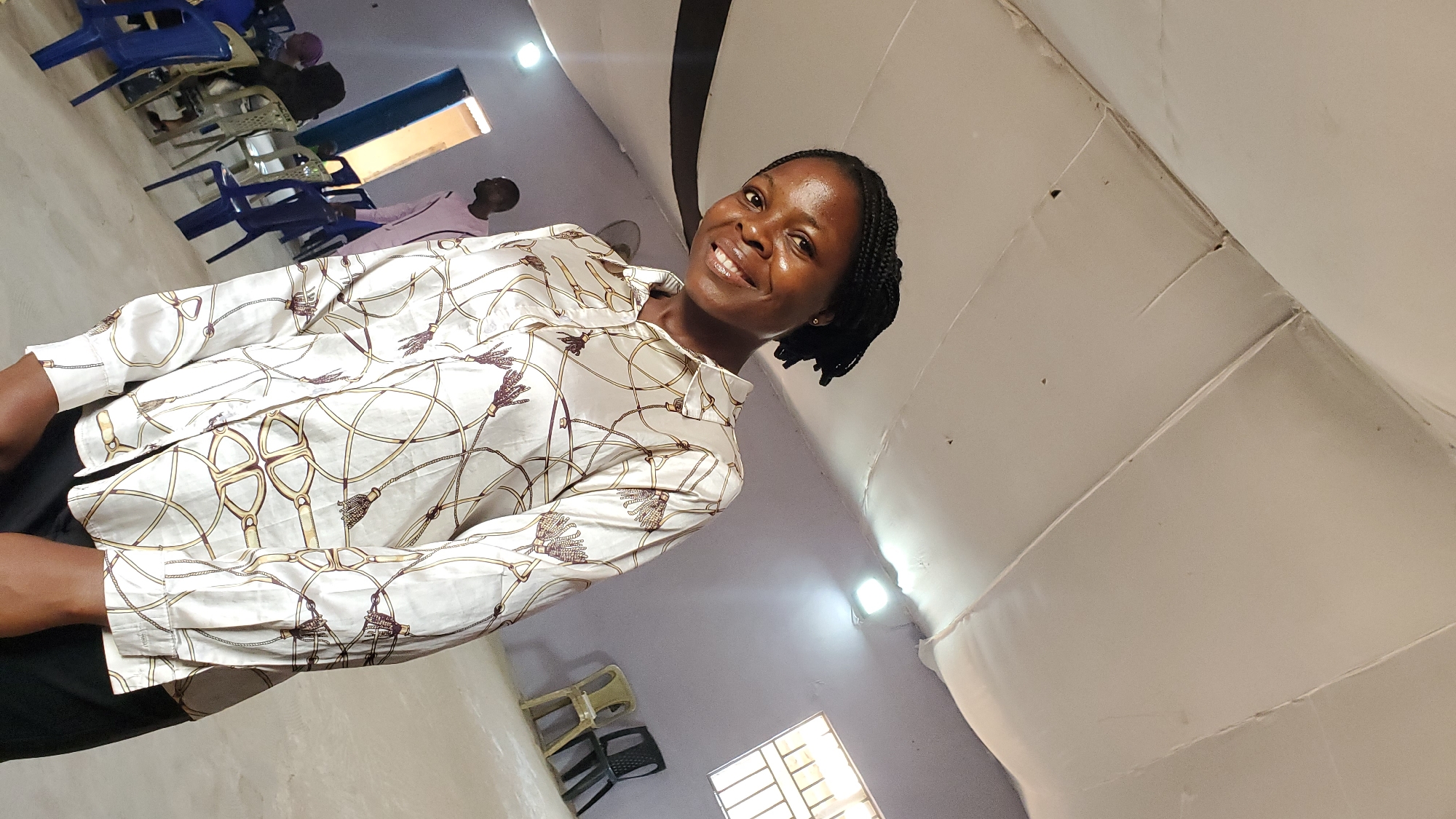 The image shows a woman in a white, patterned blouse standing in a room that appears to be a community hall or meeting space. The room has a high ceiling with exposed white panels and visible lighting fixtures. The walls are painted a light purple, and there is a window with metal bars on the right side. In the background, several chairs are visible, including blue plastic chairs and some darker, more solid chairs. Two men and one other person, whose gender is not clearly discernible, are partially visible in the background. The woman is smiling and looking towards the camera. The overall lighting suggests an indoor setting, possibly during daylight hours given the brightness. The presence of chairs and the spacious layout suggest a place designed for gatherings. The context of Jalingo, Nigeria, would imply this is a location within that city, likely a public or community building.