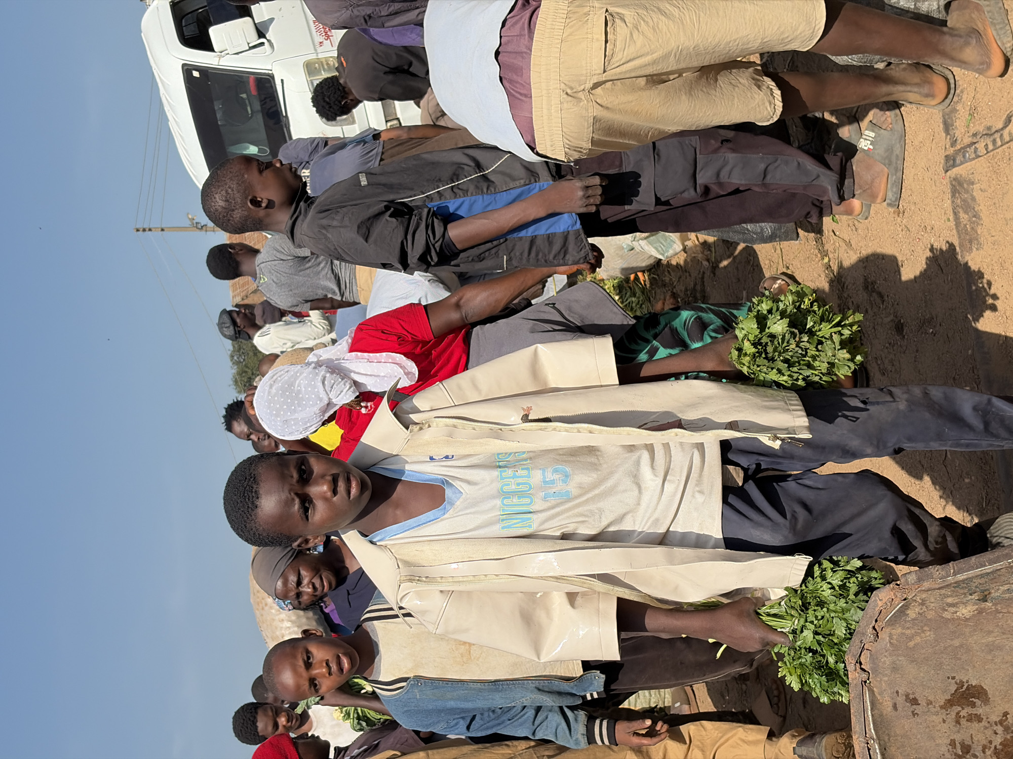 The image depicts a bustling outdoor market scene in Gura Top, Nigeria. The foreground is dominated by a young boy wearing a light-colored jacket over a white t-shirt with blue lettering and the number "15." He is holding bunches of fresh green herbs. To his left, another young boy is visible, wearing a denim jacket. A group of people, including women and men of various ages, are gathered in the background, some carrying produce and others appearing to be shoppers or vendors. A white van is parked in the upper right portion of the image. The ground is covered in dirt, with some debris and sacks visible. The overall atmosphere suggests a busy commercial exchange.