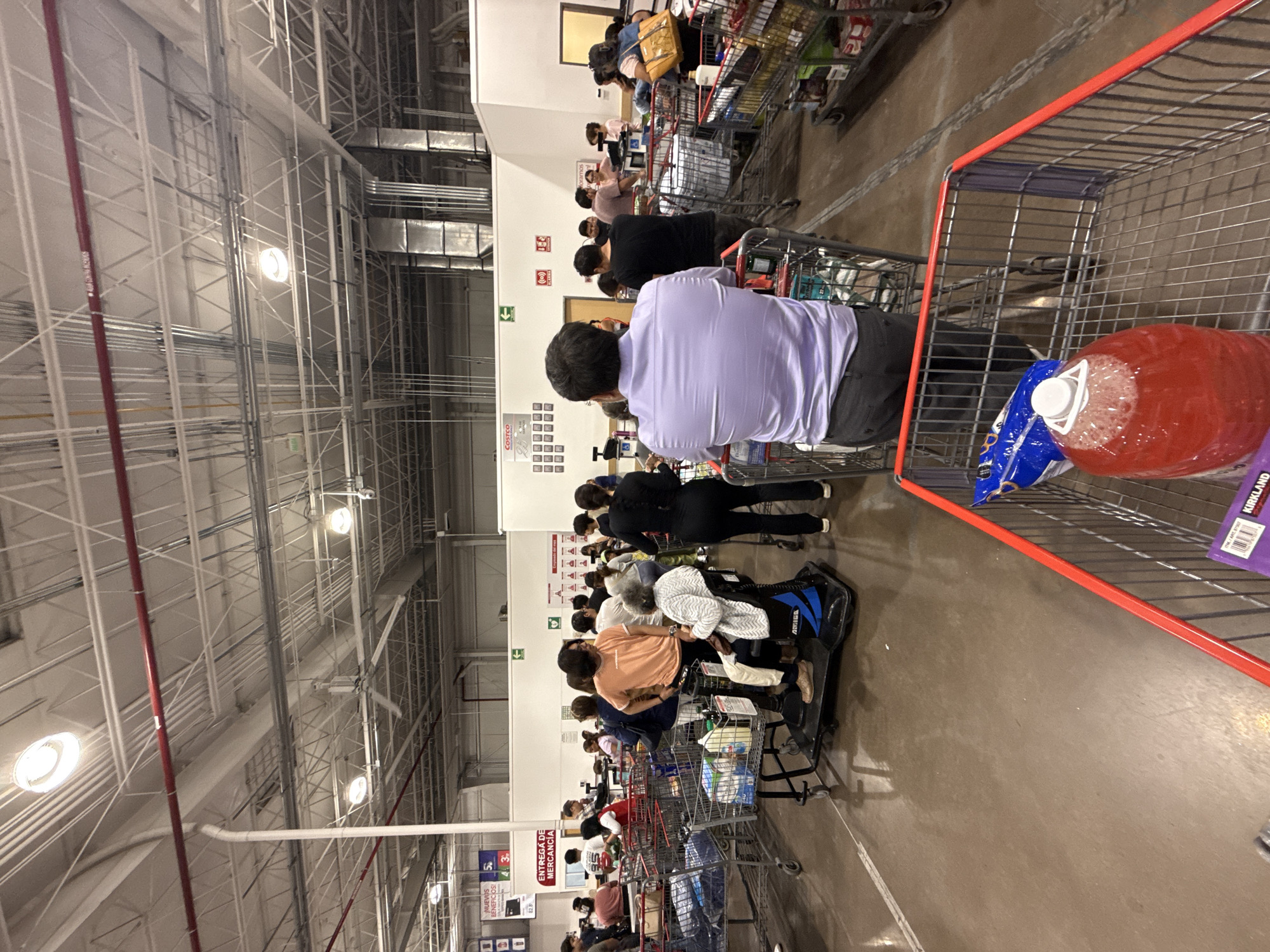 The image captures a long checkout line at what appears to be a Costco warehouse store. People are waiting with their shopping carts, their contents visible. The scene is inside a large, warehouse-style building with high ceilings and visible structural support. Signage in Spanish is present, including one referencing 'Costco' and another indicating 'Entrega de Mercancia' (Delivery of Merchandise). One item in the foreground cart is an orange liquid. The scene seems to be in Puebla, Mexico.