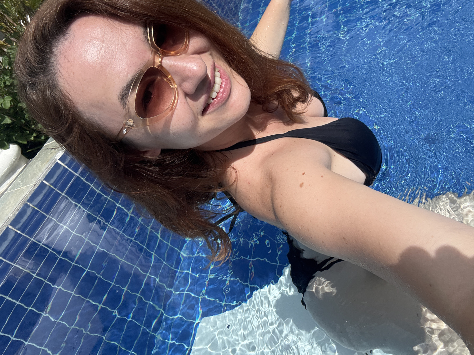 A woman with auburn hair, wearing sunglasses and a black bikini, is partially submerged in a swimming pool. She is taking a selfie, with her head tilted back and a wide smile on her face. The water is a vibrant blue, and the sunlight creates shimmering patterns on the surface. The pool has tiled walls, visible at the bottom left of the image. The overall setting appears to be a resort or private residence, with lush green foliage visible in the upper left corner. The location is specified as Baan Taling Ngam, Thailand.