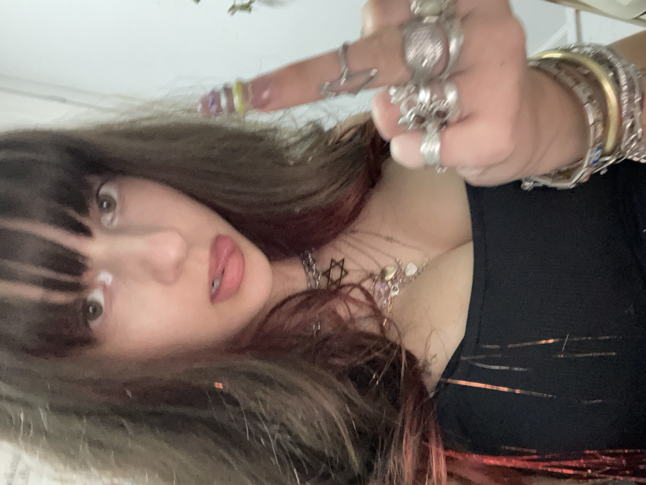 A woman with brown hair and bangs, a fair complexion, and full pink lips poses at a low angle, wearing a black top. She’s adorned with numerous rings and bracelets, including one with a skull design. She also wears a necklace featuring a Star of David pendant. Her middle finger is extended, while the others are curled into a fist. The background is a blur of light and color, possibly from decorations.