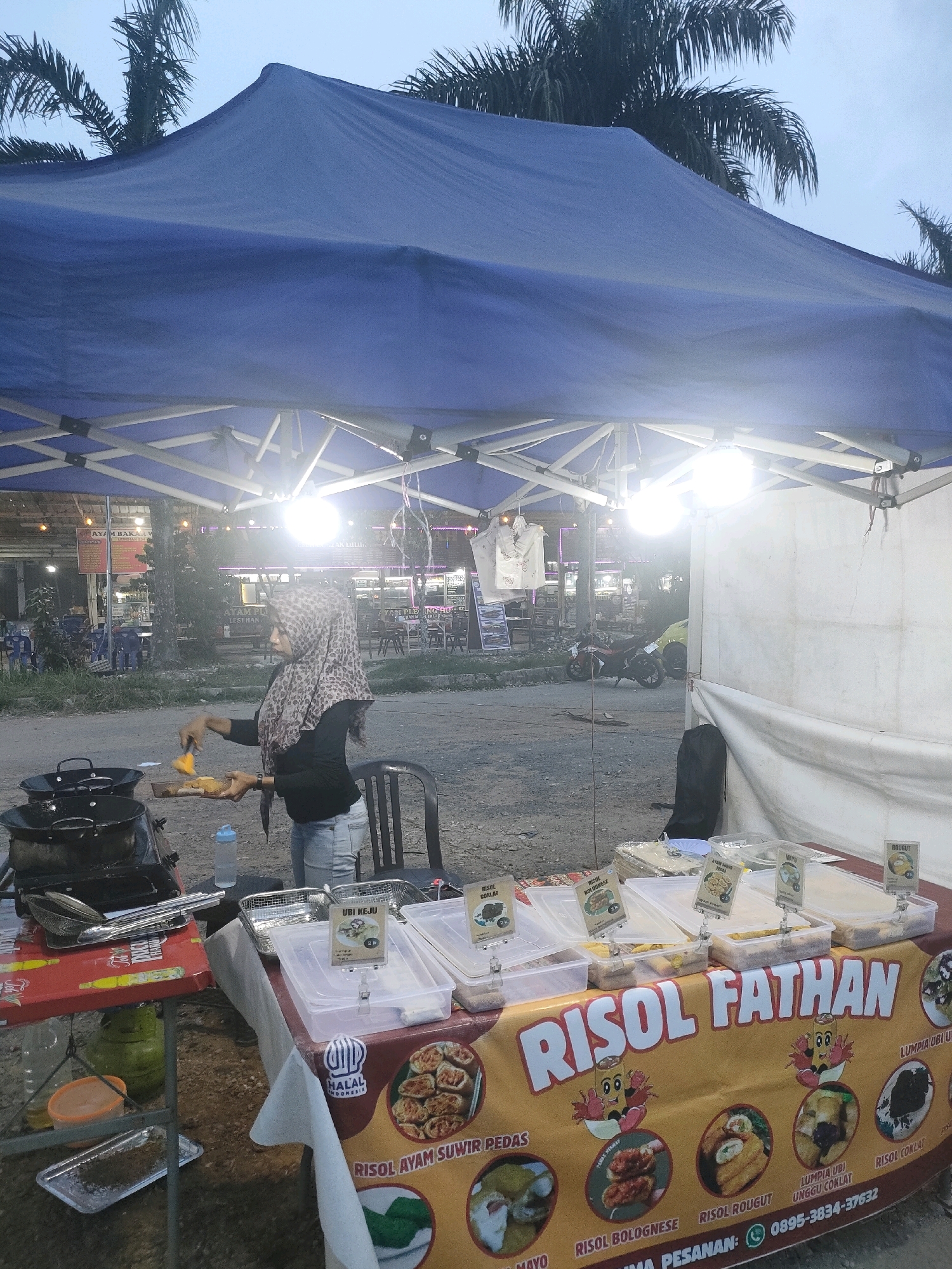 In Batam, Indonesia, an outdoor food stall is set up under a blue tent. A woman wearing a hijab and a black long-sleeved shirt over light-colored jeans is standing on the left side of the frame, cooking. She is holding a pan and a spatula, appearing to be preparing food. In front of her, on a table, are two black woks on a stove, a bottle of water, and various cooking utensils.
The stall displays several plastic containers filled with food items. A banner across the front of the table reads "RISOL FATHAN" and features illustrations of food. Various labels with names like "RISOL AYAM SUWIR PEDAS", "RISOL BOLOGNESE", "RISOL ROGUT", and "LUMPIA UBI UNGGUL COKLAT" are visible on the food containers. The background shows other stalls and structures, suggesting a marketplace or roadside food area. Palm trees are visible in the upper part of the image, indicating a tropical environment. The lighting suggests it is either early morning or late evening.
