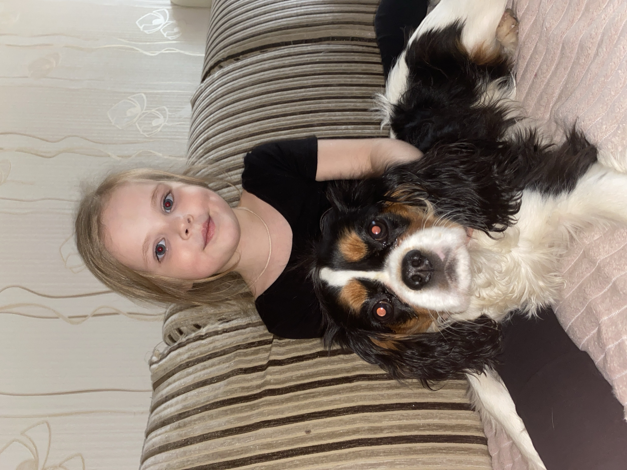 An eye-level indoor shot, taken in Votkinsk, Russia, captures a young girl and a tri-color dog lying together on a striped couch. The camera's flash has caused a red-eye effect in both subjects' eyes.
On the left, the young girl, with blonde hair and blue eyes, looks towards the camera with a gentle smile. She is wearing a black short-sleeved top and a thin, light-colored necklace. Her left arm is visible, resting around the dog, indicating a close and affectionate interaction.
To the right, the dog, a small to medium-sized breed with black, white, and tan markings, is also looking directly at the camera. Its fur, particularly around the face, appears somewhat wet or matted. The dog is positioned close to the girl, with its head centered in the frame.
Both subjects are lying on a couch upholstered with a pattern of alternating beige and brown stripes. In the upper left background, a light-colored wall with subtle, wavy or floral patterns is visible. The scene suggests a calm moment of relaxation or play between the child and her pet in a home environment. There is no visible text in the image.