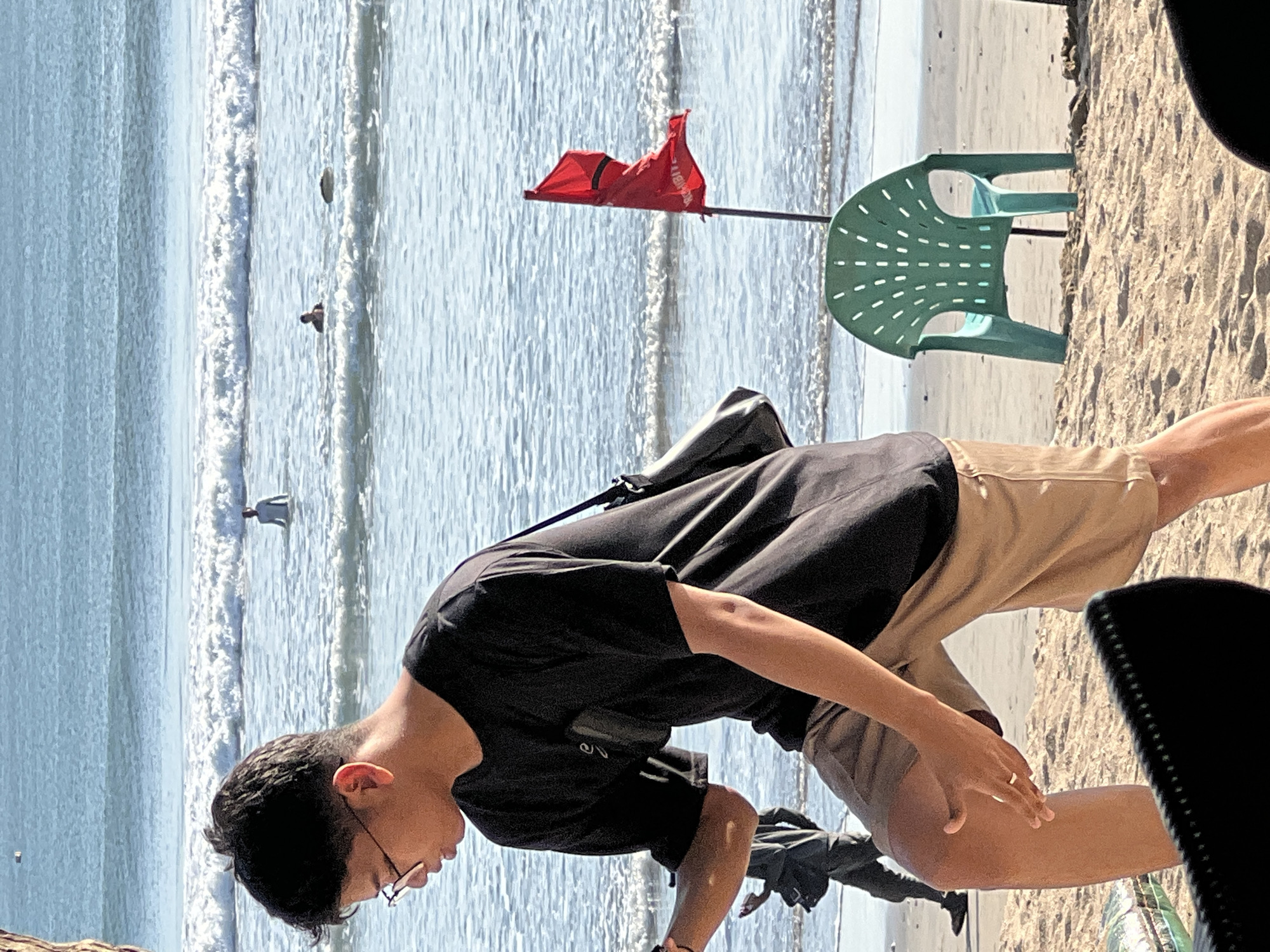 A young man wearing glasses, a black t-shirt, and khaki shorts walks on the sandy beach of Kuta, Indonesia. He carries a dark shoulder bag. In the background, waves roll onto the shore, and several people are visible in the water, one standing and two in the distance. A red flag, likely indicating a swimming hazard, stands planted in the sand near a green plastic chair. The scene is bathed in sunlight, suggesting it is daytime.