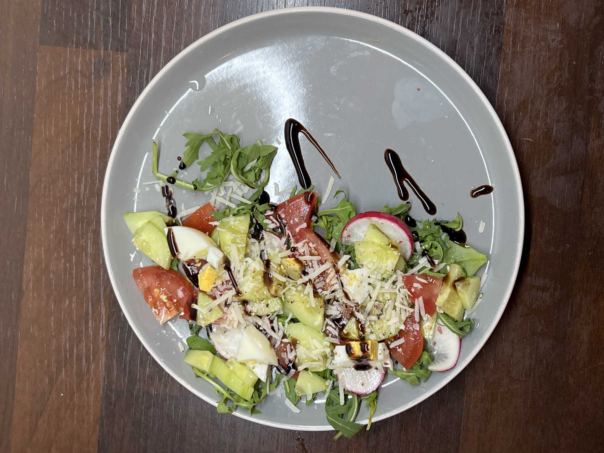 A close-up, overhead shot showcases a grey plate of salad placed on a dark wooden table. The salad is composed of arugula, sliced radishes, cherry tomatoes, cucumber, hard-boiled egg, and shredded parmesan cheese. A balsamic glaze is drizzled over the salad and onto the plate. The scene is set in Balbriggan, Ireland.