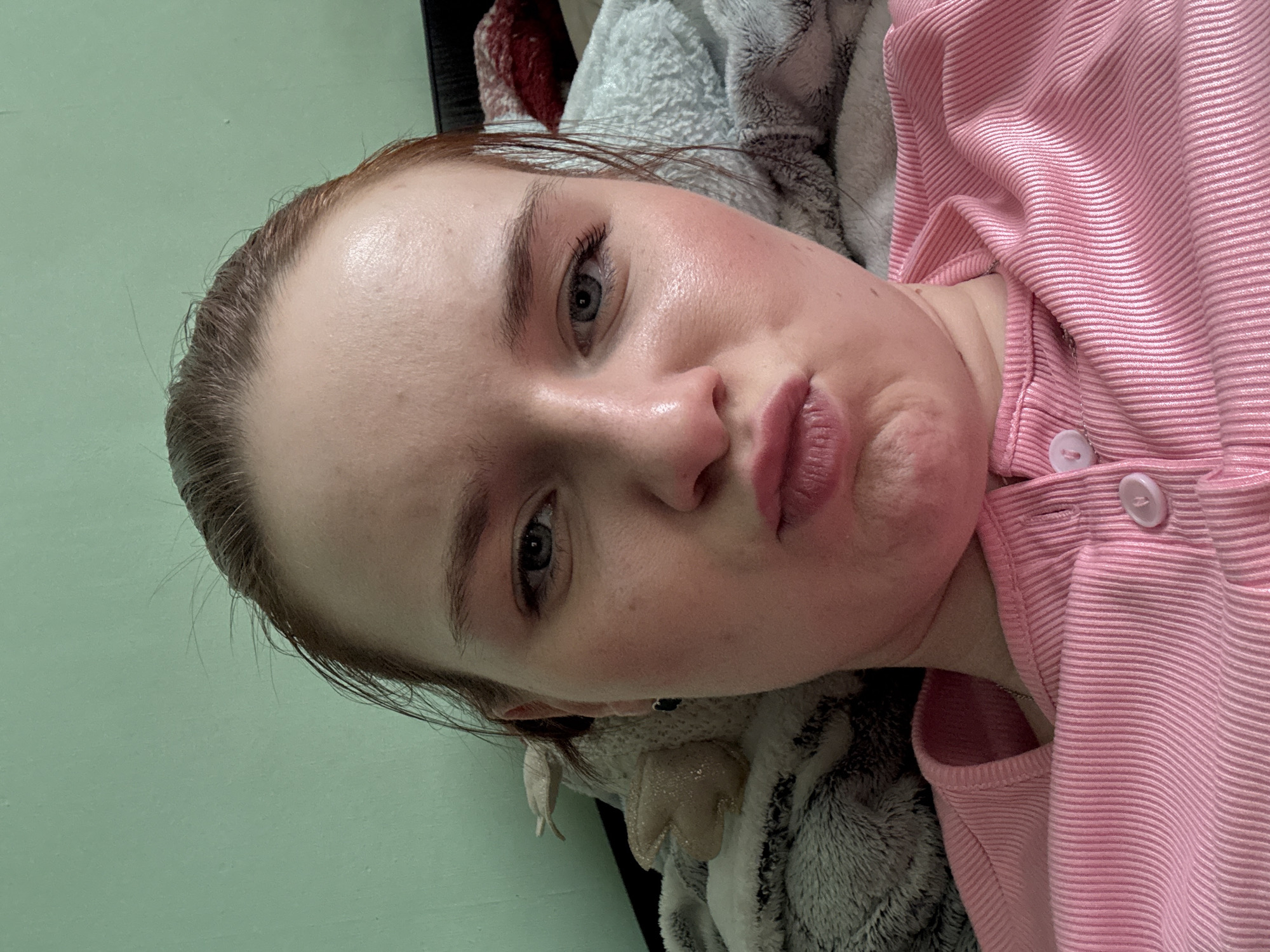 The image shows a close-up portrait of a fair-skinned individual with light brown hair. The person is lying down, with their head tilted slightly to the right. They have light-colored eyes and pursed lips. They are wearing a pink, button-down shirt made of ribbed fabric. The background includes a light green wall and a gray and white textured blanket or throw. The location and city in Russia are unknown.
