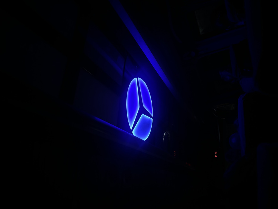 The scene depicts the interior of a darkened space, located in the city of Khashuri, Georgia. A prominent blue, oval-shaped light fixture, divided into three illuminated segments, is mounted on the ceiling in the upper center of the frame. This fixture is the primary light source, casting a deep blue hue across the visible ceiling panels and structural elements. The majority of the lower and peripheral areas of the image remain in profound darkness due to the low light conditions. In the lower-left quadrant, a faint red digital display indicates "04:16," with additional small red indicator lights visible above it. Indistinct dark forms in the lower foreground suggest the presence of occupied seats or individuals within the dimly lit environment.