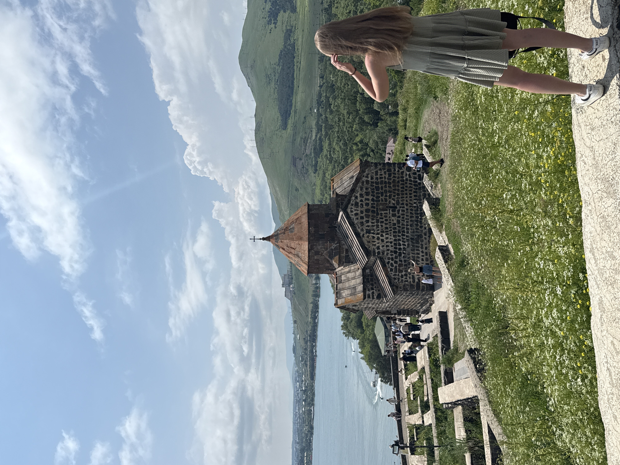 An outdoor, eye-level shot captures a scenic view of the Sevanavank monastery complex overlooking Lake Sevan in Sevan, Armenia. In the foreground, a young woman with long blonde hair, wearing a light green ruffled dress and white Nike sneakers, stands facing away from the camera, her hands raised to her head as if shielding her eyes from the sun. She is positioned on a stone ledge adorned with a field of white wildflowers and green grass. Behind her, the ancient stone monastery, with its characteristic conical roof and cross, is prominently featured.
Below the monastery, a body of water, Lake Sevan, stretches out, with a few boats visible on its surface and a town or city nestled along its far shore. The background consists of lush green, rolling hills and mountains under a partly cloudy sky with a bright sun. Several other visitors are dispersed throughout the complex, walking along pathways and observing the historical site. The overall atmosphere is serene and picturesque, showcasing the natural beauty and historical significance of the location.