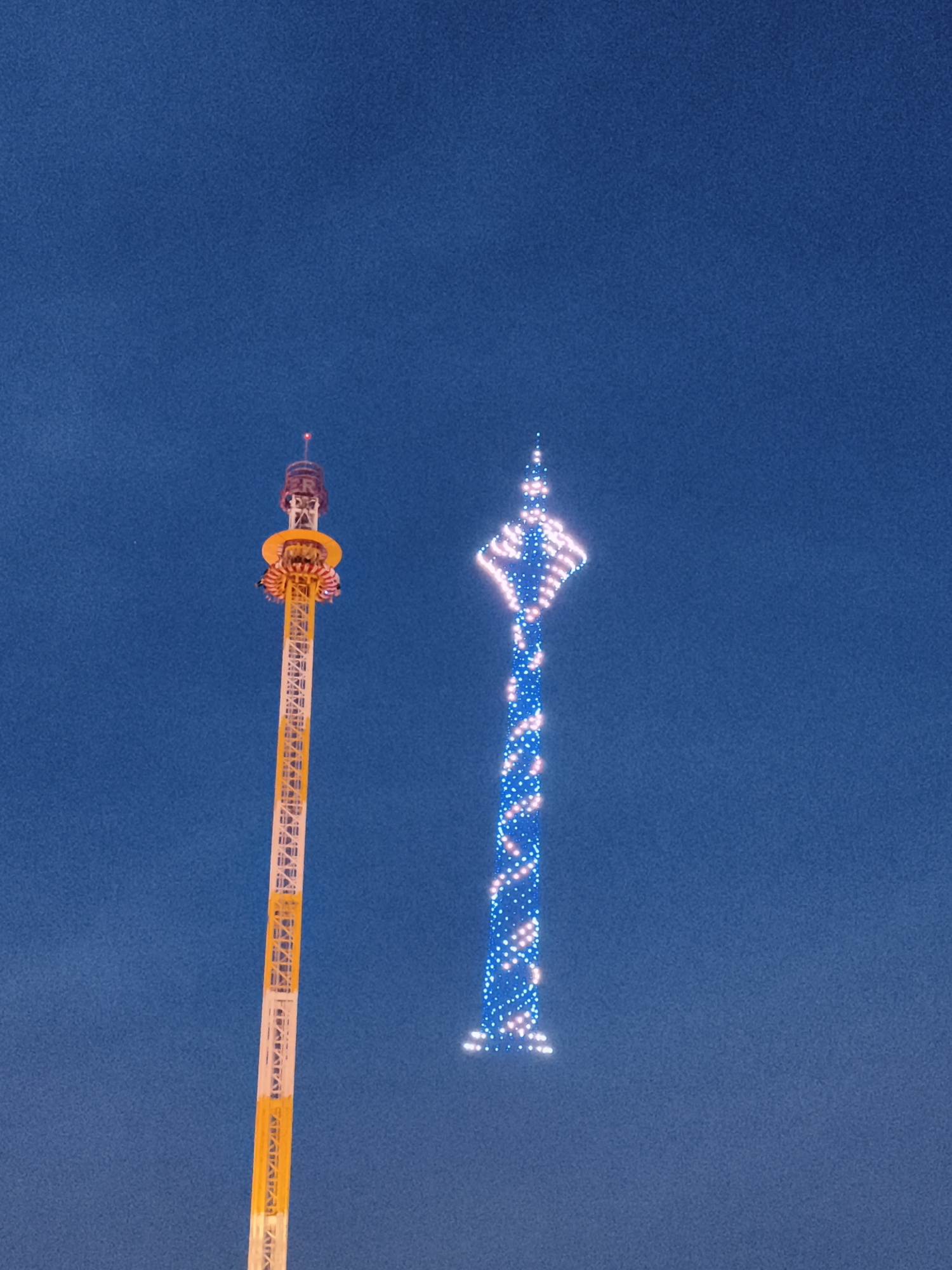 The image shows two illuminated towers against a deep blue sky, likely taken at dusk or night. On the left is a tall, yellow and white lattice tower, part of a drop tower amusement ride, with a circular platform visible near the top. Several people are seated on this platform, likely about to experience the ride. On the right is a slender, tapering tower adorned with a spiral pattern of blue and white lights. This tower is likely the Rhine Tower (Rheinturm) in Dusseldorf, Germany, as evidenced by its distinctive shape and illumination. The sky is clear and uniform in color.