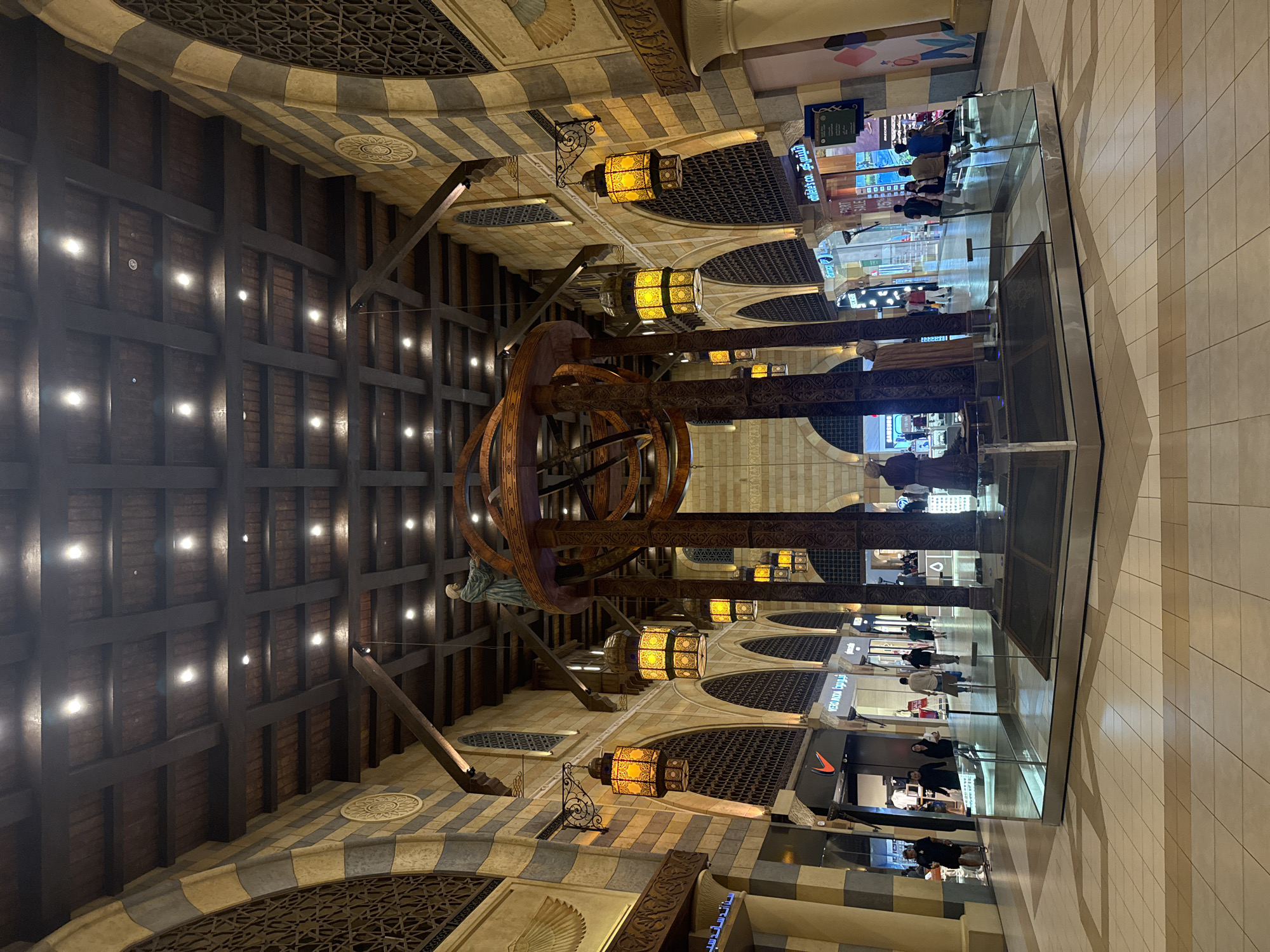 The image captures an interior view of the Souk Madinat Jumeirah in Dubai, United Arab Emirates, a popular marketplace designed to resemble a traditional Arabian souk. In the center of the frame, a large, elaborate wooden structure, possibly an astrolabe or a celestial globe, dominates the scene. This central installation is adorned with what appear to be two statues or mannequins dressed in traditional Middle Eastern attire. The structure is lit by several ornate, yellow glass lanterns hanging from the ceiling.
The architecture features high, arched ceilings with dark wooden beams and panels, interspersed with circular decorative elements. The walls are constructed with alternating bands of light and dark stone or plaster, giving it a textured, aged appearance. Large, intricately patterned arched windows or screens are visible along the walls, adding to the traditional souk ambiance.
The foreground shows a polished tiled floor with a geometric pattern. A modern glass and metal barrier surrounds the central installation, contrasting with the more traditional surroundings. Beyond the central exhibit, a bustling shopping area is visible, with various retail stores lining the walkway. Signs for brands like Nike and Skechers can be observed. Several individuals, dressed in casual contemporary clothing, are seen walking through the mall, browsing the shops, or standing in small groups. The lighting in the mall appears to be a mix of ambient overhead lights and the warm glow from the decorative lanterns. The overall impression is a blend of traditional Middle Eastern architecture and modern retail commerce.