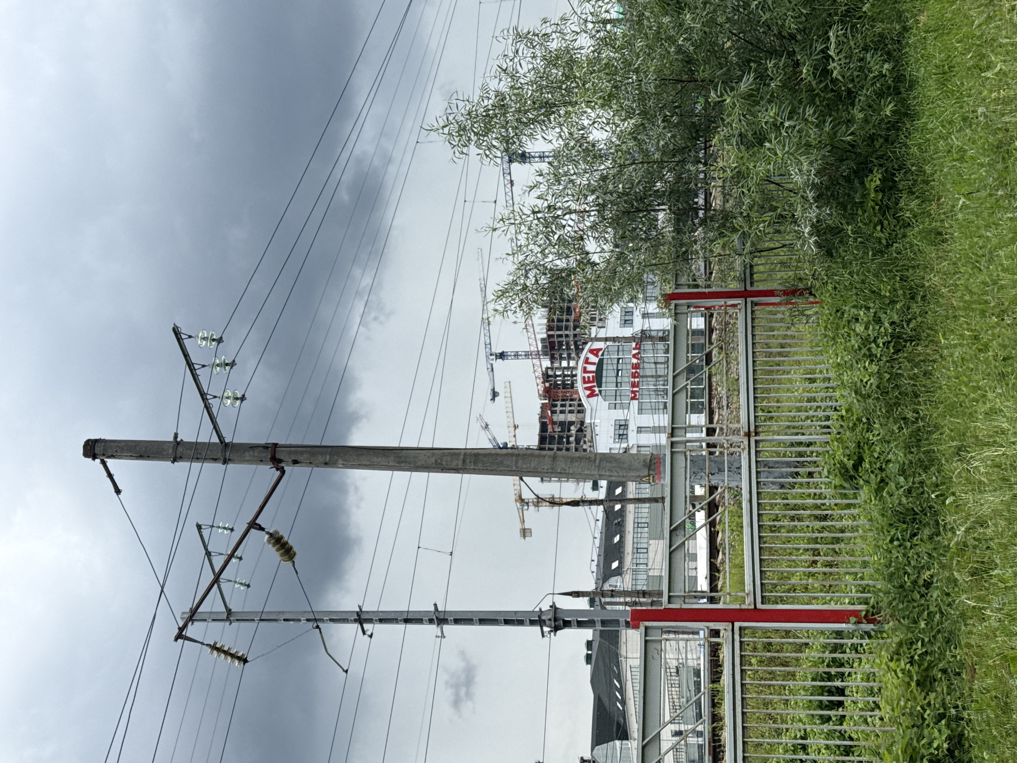 This image captures an outdoor scene with multiple subjects and objects. A prominent element is a utility pole with electrical wires stretching across the frame, suggesting a connection to a power grid. In the background, a large building is under construction, with several cranes visible, indicating ongoing development or infrastructure work. The building itself appears to be modern, with a white facade and large windows. A prominent red and white sign is visible on the building, with text that reads "МЕГГА".
To the right of the image, a metal fence with red accents surrounds an area with lush green grass and vegetation. This fence separates the viewer's perspective from the construction site and the grassy area, creating a sense of boundary. A tree with dense foliage is situated between the utility pole and the building, partially obscuring the view of the construction.
The sky is overcast and gray, suggesting a cloudy or potentially rainy day. The lighting is diffused, and there are no strong shadows. The overall mood of the image is industrial and somewhat stark, due to the construction and the utility infrastructure, contrasted with the natural element of the grass and tree. The orientation of the image is vertical, with the ground appearing at the top and the sky at the bottom. The exact time of day is difficult to determine due to the overcast conditions. The location is unknown, but the presence of construction and utility infrastructure points towards an urban or suburban environment. No people are visible in the image.