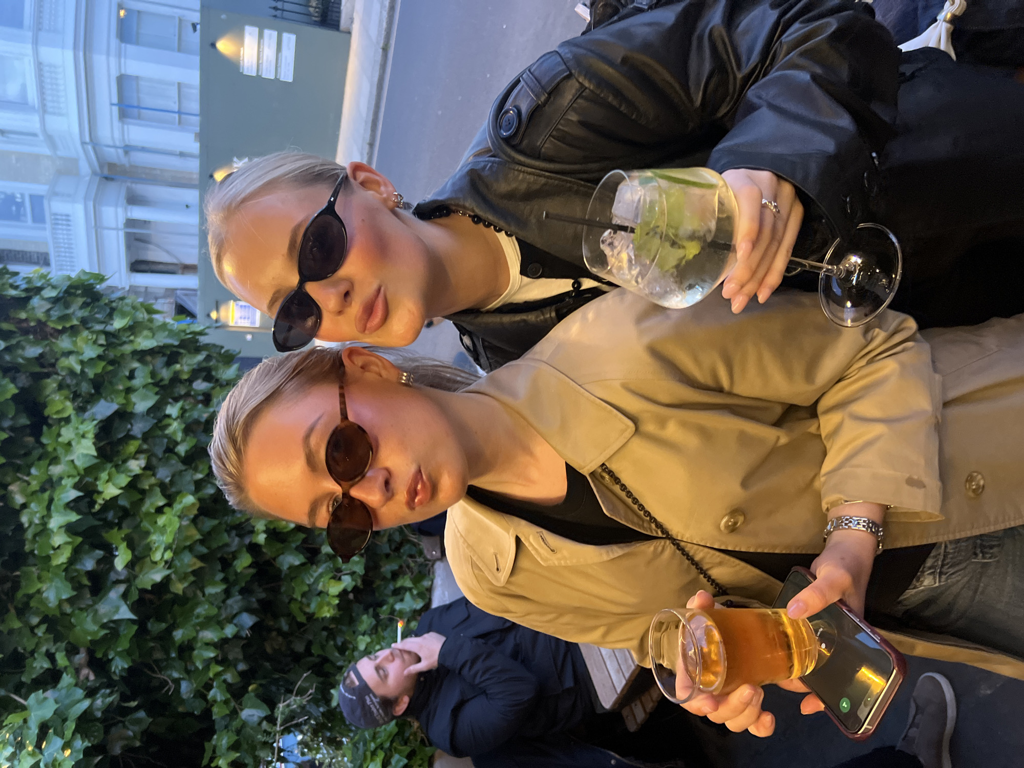 Two blonde women wearing sunglasses are posing for a photograph while holding drinks. The woman in the foreground is wearing a tan trench coat and holding a shot of beer and a mobile phone, while the woman in the background is wearing a black leather jacket and holding a cocktail. Both women are wearing jewelry. A man wearing a black hat and holding a cigarette is visible behind the woman in the foreground. The background includes a building and green foliage. Based on the architecture and general ambiance, the location is likely in a public area of London, United Kingdom.