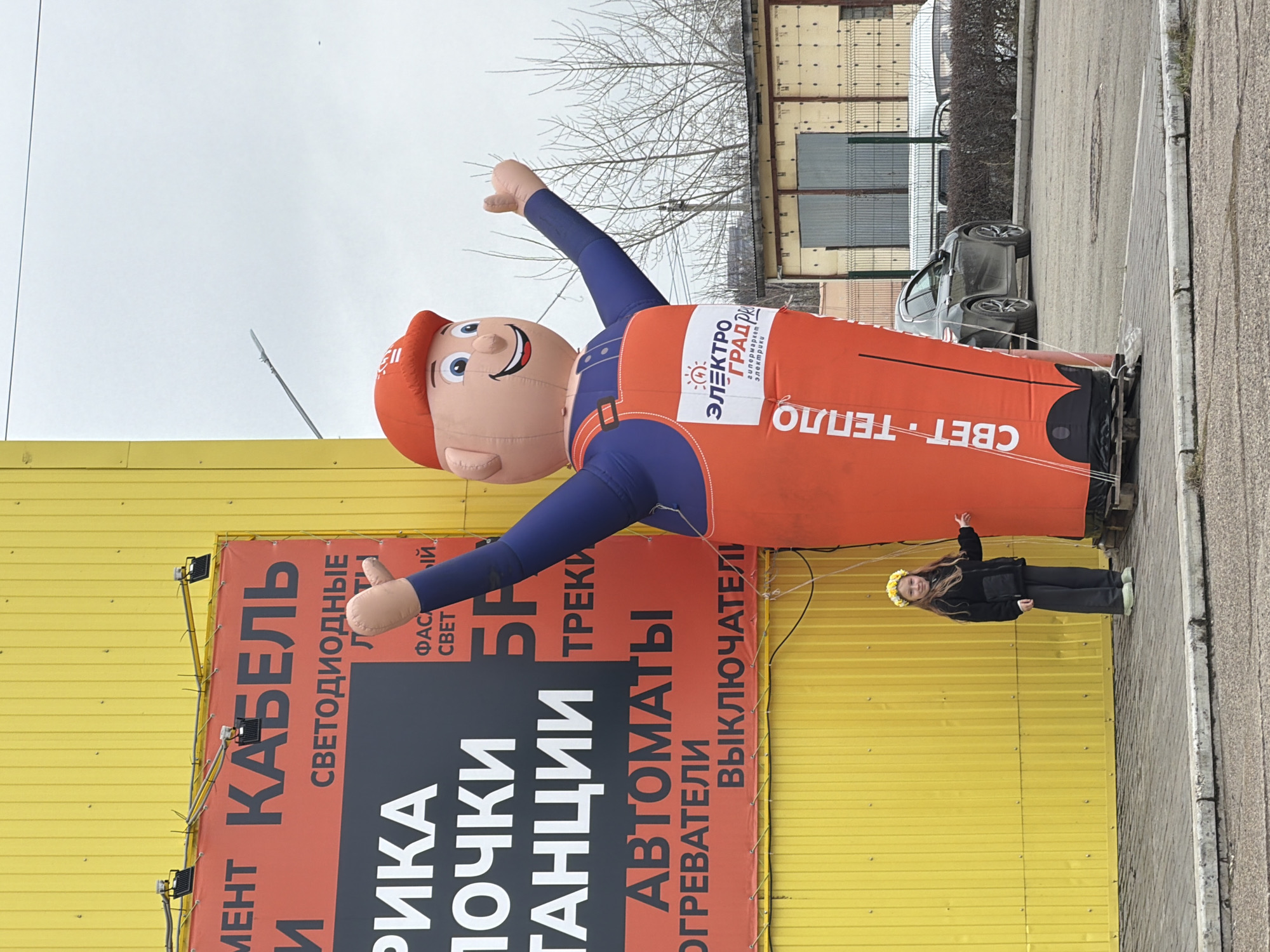 This image captures an outdoor scene in Chaikovsky, Russia, depicting a large inflatable mascot and a young girl standing in front of a commercial building. The setting is likely a street or parking lot, with an overcast sky suggesting a cool daytime environment.
The main subject is a tall, smiling inflatable mascot, designed as a cartoon man in a red cap, blue long-sleeved shirt, and orange overalls. It has one arm raised and the other pointing downwards. A logo on its chest reads "ЭЛЕКТРО ГРАД РА" (Elektro Grad Ra) and "гипермаркет электрики" (hypermarket of electrical goods), indicating it promotes an electrical supplies store. The phrase "СВЕТ ТЕПЛО" (Light Heat) is also visible on its lower half.
To the right of the mascot, a young girl with long brown hair stands, looking towards the camera with a slight smile. She wears a dark jacket, dark pants, light green shoes, and a distinctive yellow floral wreath on her head.
The background features a building with a bright yellow corrugated metal facade. A prominent red and black sign with white text is mounted on the building. The visible text lists various products, including "КАБЕЛЬ" (cable), "СВЕТОДИОДНЫЕ" (LED), "ТРЕКИ" (tracks/spotlights), "АВТОМАТЫ" (automatic circuit breakers), "ОГРЕВАТЕЛИ" (heaters), and "ВЫКЛЮЧАТЕЛИ" (switches), reinforcing the theme of electrical goods. Other partial words like "ФАСА CBET" (Facade Light), "РИКА", "ПОЧКИ" (buds/kidneys), and "ГАНЦИИ" are also present.
In the distance, bare trees are visible under the gray sky, and several cars are parked along the street, typical of an urban or suburban commercial area.