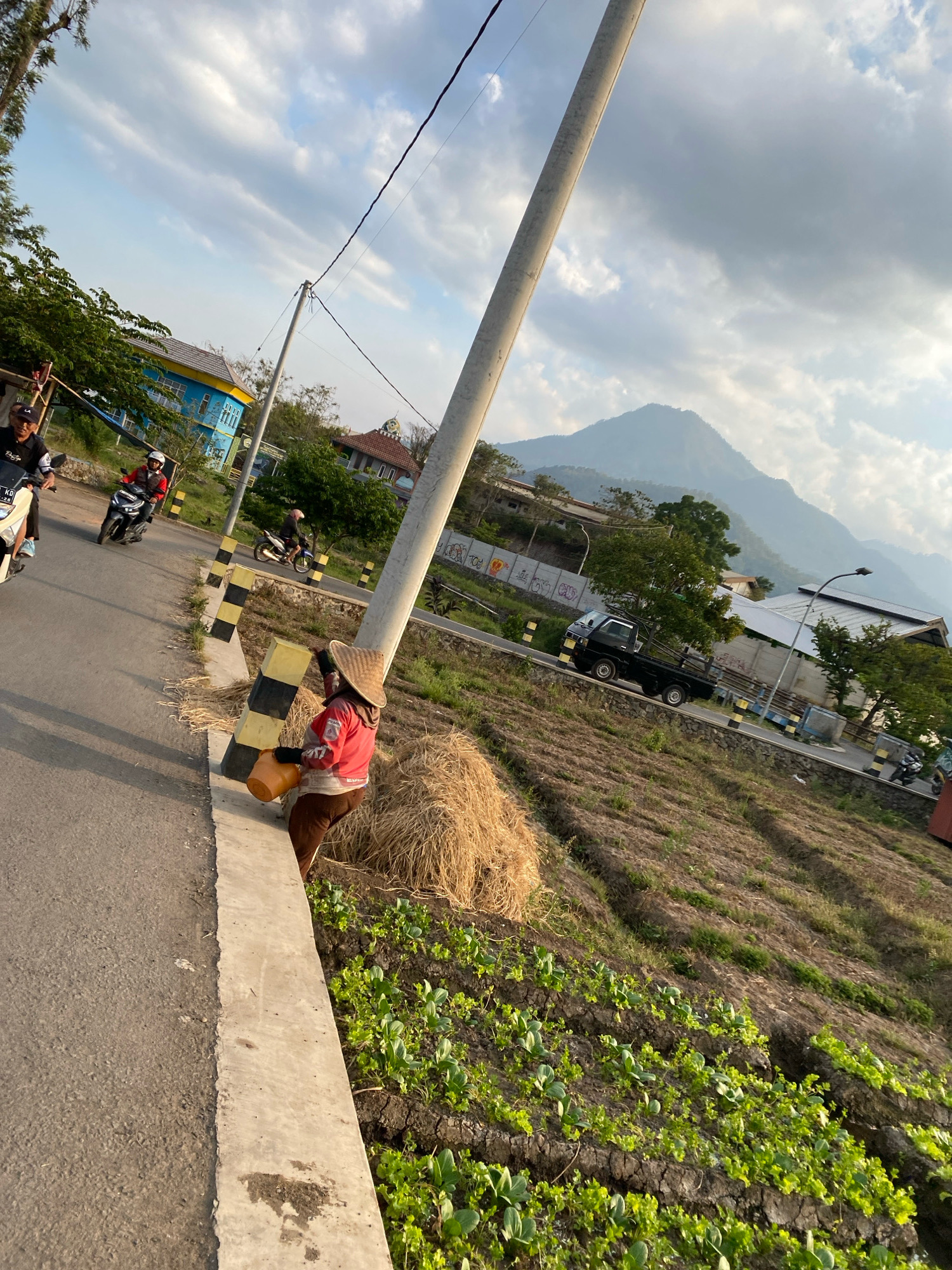 The image captures a rural roadside scene under a cloudy sky. A person wearing a conical hat, a red jacket, and brown pants is standing on the side of a paved road, holding an orange bucket. The road has yellow and black striped barriers along its edge. Several motorcycles are visible on the road, with some riders wearing helmets. In the background, there are buildings with blue and yellow facades, trees, and a prominent mountain in the distance. Adjacent to the road, a field with rows of crops is visible. A black pickup truck is also seen on the road. The lighting suggests it is either morning or late afternoon.