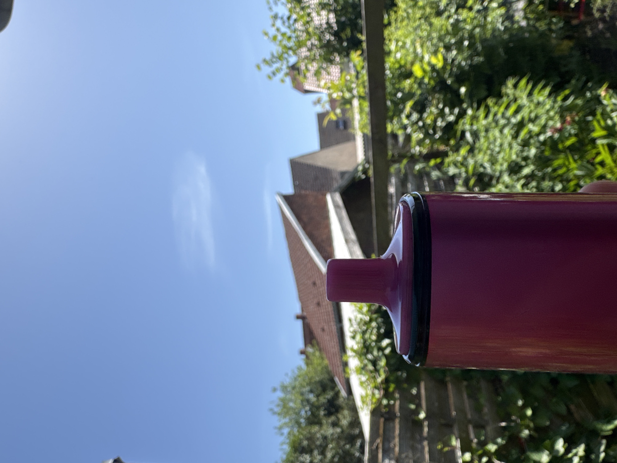 The image displays a close-up of a maroon-colored cylindrical object, possibly a travel mug or a reusable water bottle, in the foreground. The object has a black rim and a small, protruding section also in maroon. The scene is outdoors, with a bright blue sky dominating the upper left portion of the frame, suggesting it is daytime, likely sunny.
In the background, out of focus, are elements of a residential setting. A red-tiled roof of a building is visible, along with some light-colored walls and windows. Green foliage, likely trees and bushes, surrounds the building and creates a lush backdrop. A wooden fence can also be seen behind the main object. The overall impression is one of a casual, everyday outdoor environment on a clear day. No people or specific events are depicted, and there is no visible text in the image. The focus is primarily on the object in the foreground, with the background providing context for its placement.
