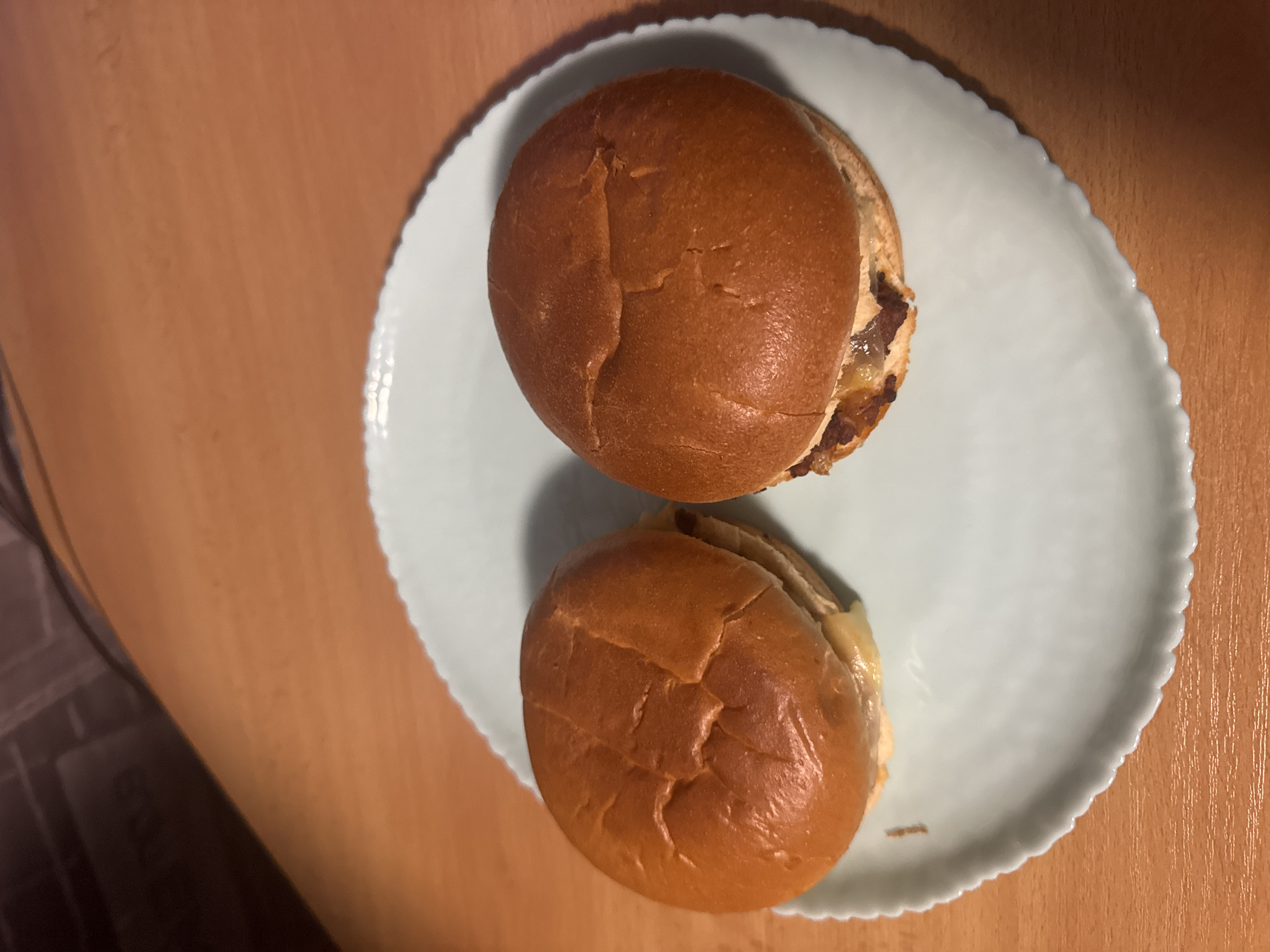 Two burgers, likely cheeseburgers, are presented on a light blue or mint green plate with a decorative scalloped edge. Both burgers feature golden brown, slightly shiny buns with visible creases and cracks. Melted cheese and what appears to be browned meat or caramelized onions are visible peeking out from under the top buns.
The plate rests on a warm-toned, wooden surface, suggesting an indoor setting such as a table or desk in Sofia, Bulgaria. The lighting appears to be artificial or diffused, casting soft shadows under the burgers and the edge of the plate. A darker, indistinct area is visible in the background on the left side of the frame.
No people are visible, and the scene depicts no specific activity other than the static presentation of the food, ready for consumption. There are no distinctive location cues within the image itself beyond the provided context of Sofia, Bulgaria, and no visible text.