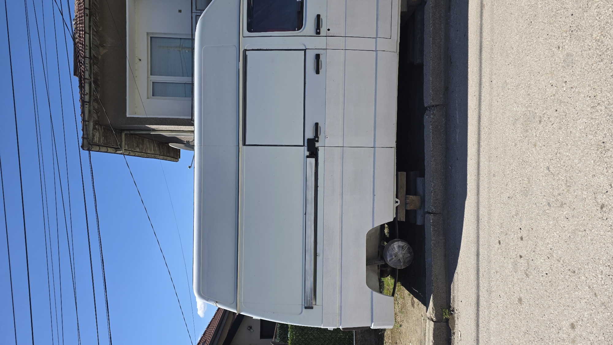 The image depicts the side of a white van parked on a street. The van is positioned adjacent to a sidewalk and curb. Behind the van, there's a building with a window and a tiled roof. Overhead, power lines are visible against a clear blue sky. The location is estimated to be in an urban area, in the city of Blagoevgrad, Bulgaria.