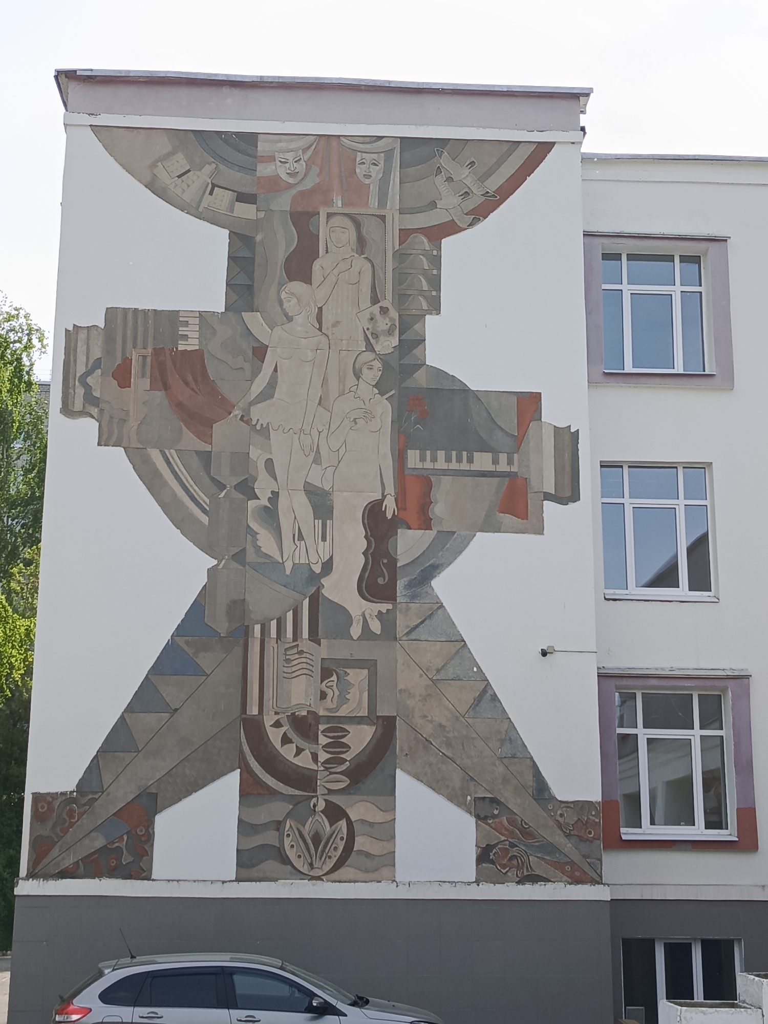 The image shows a large, artistic mural painted on the side of a building. The mural features stylized figures of women and various objects related to the arts, such as musical instruments, theater masks, and books. The style is somewhat abstract and geometric, with sections filled with triangles, circles, and waves. The overall color palette is muted, with grays, browns, and hints of red and blue.
The building itself is three stories tall and has several windows on the side adjacent to the mural. The windows have white frames, and the building is painted white, except for the gray base. A silver car is partially visible at the bottom of the frame.
The image is taken in the city of Chaikovsky, Russia.
