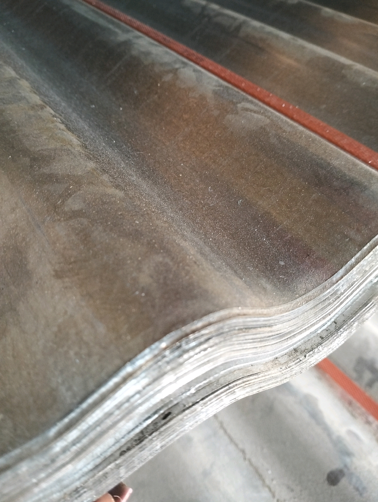 The image displays a stack of corrugated roofing sheets, possibly made of asbestos cement, with a reddish band running diagonally across the upper portion. The sheets are weathered and dusty, showing signs of age and exposure to the elements. The perspective is a close-up, focusing on the texture and layered structure of the material. The background is blurred, but appears to be a concrete surface. There is no specific identifiable location within the city of Kinanie ward, Kenya, visible in the image. The scene is static and depicts an accumulation of building materials.