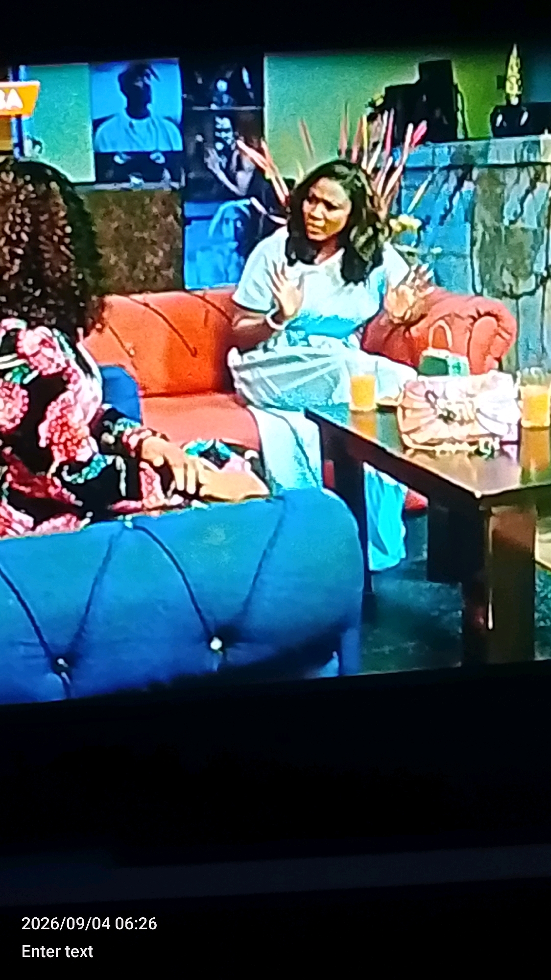 The image depicts an indoor scene with two women on a couch, possibly in a studio or living room setting. The woman on the right is wearing a light blue outfit and has her hands raised, appearing to be speaking or gesturing. She is seated on an orange couch. To her left, another woman with curly hair, wearing a floral-patterned top, is partially visible on a blue couch. A wooden table is situated in front of the couch, with two glasses of orange liquid and a patterned clutch purse on it. In the background, there is a wall with framed images and decorative elements. The overall ambiance suggests a conversation or interview setting. The provided timestamp at the bottom indicates it was captured on September 4, 2026, at 06:26. There is no visible indication of the city of Lokoja, Nigeria, within the image itself.