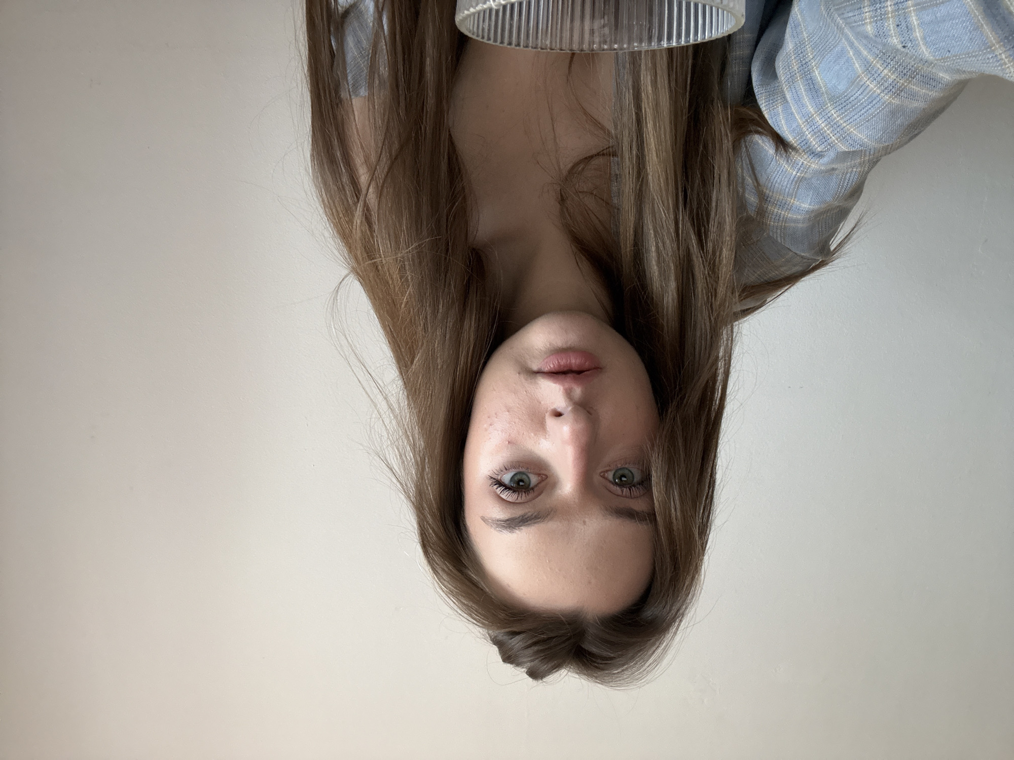 An upside-down photograph features a young woman with long brown hair. Her face is centrally positioned. She has fair skin, green eyes, and slightly parted pink lips. Above her head is a blurred, ribbed cylindrical object. A blue and white plaid garment appears in the upper right corner of the frame. The background is a plain, light-colored wall. The location is not possible to determine from the image.