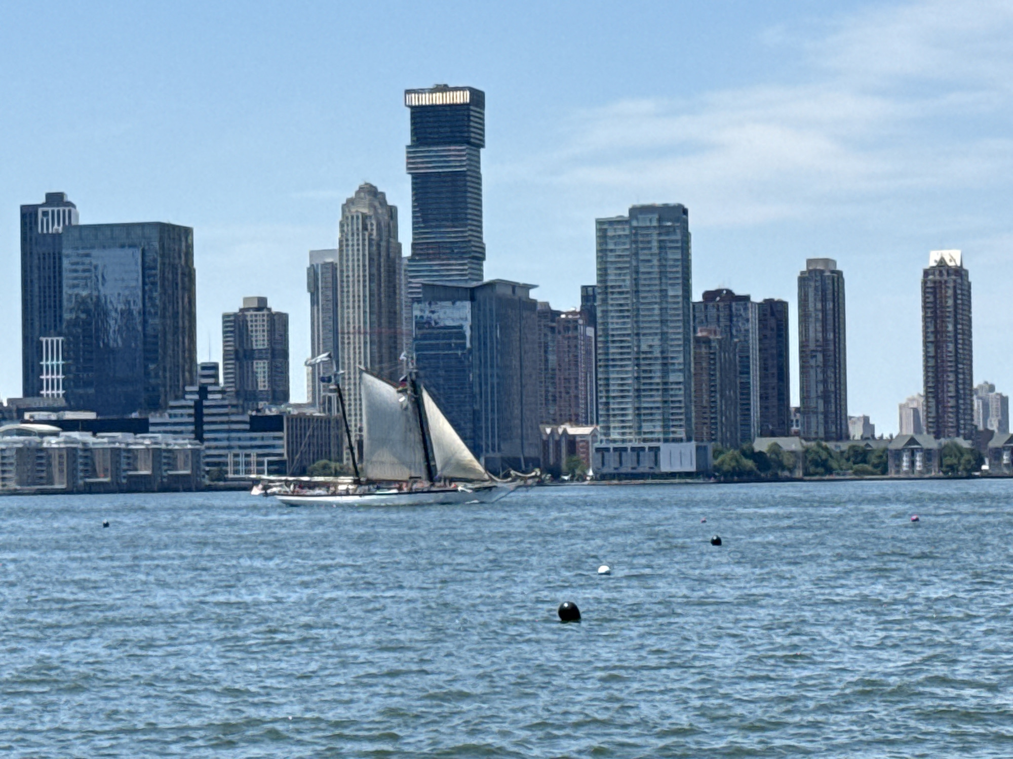 A sailboat with white sails is on the water in front of a New York City skyline. The skyline consists of numerous tall buildings of varying architectural styles, with a bright blue sky overhead. There are several buoys in the water.
