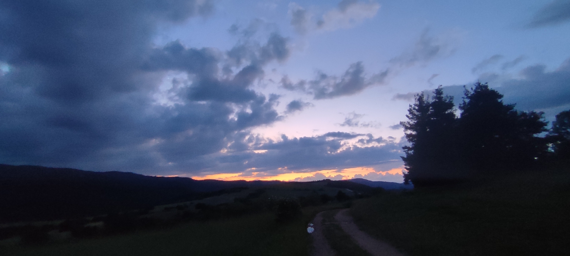 This image depicts a rural landscape at dusk in Gutsal, Bulgaria. The upper two-thirds of the frame are dominated by the sky, which transitions from dense, dark grey clouds on the far left to lighter blue and purple hues towards the horizon. A horizontal band of orange and pink light, indicative of a sunset, is visible along the distant ridge line. Various clouds, some heavily shadowed and others partially illuminated, are scattered across the sky. The lower third of the image features a dark, silhouetted mountainous or hilly terrain in the midground. In the foreground on the right, a cluster of dark, coniferous trees stands in strong silhouette against the twilight sky. A winding dirt path, lighter in tone than the surrounding shadowed vegetation, traverses the lower right quadrant of the frame. The scene is illuminated by ambient twilight, casting the terrestrial elements into deep shadow. No human figures, animals, or artificial structures are present.