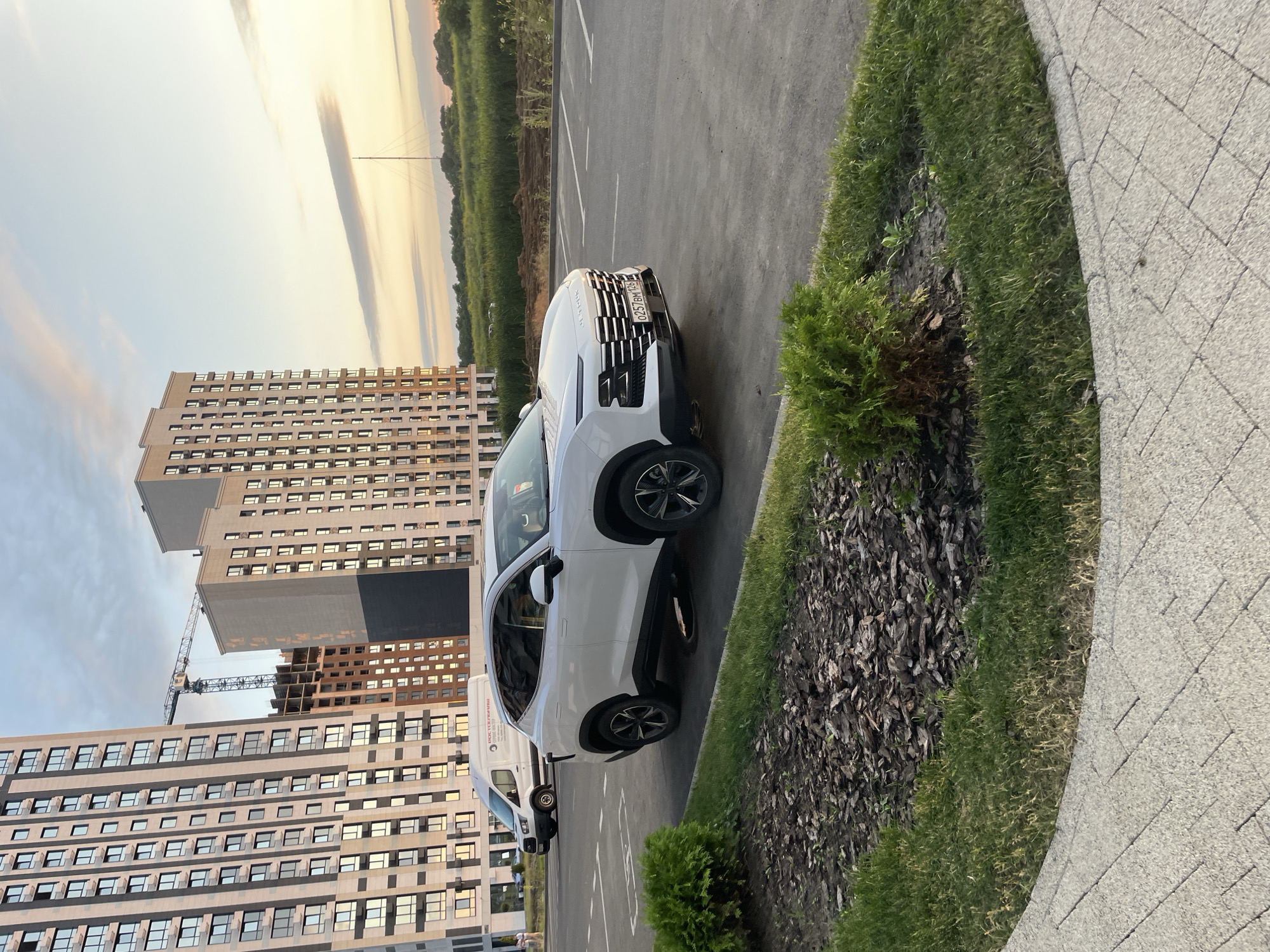 A white SUV is parked in a paved parking lot in front of a residential construction site. The SUV has a license plate that reads "O 257 BM 126". In the background, there are several multi-story apartment buildings, some of which are still under construction, and a large crane. A white van with Russian text on its side is also visible in the parking lot. The sky is partly cloudy with the sun low on the horizon, indicating either sunrise or sunset. The foreground features a patch of grass and mulch. The image appears to be taken in the city of Stavropol, Russia, as suggested by the license plate format and the architectural style of the buildings, which are characteristic of modern Russian urban development.