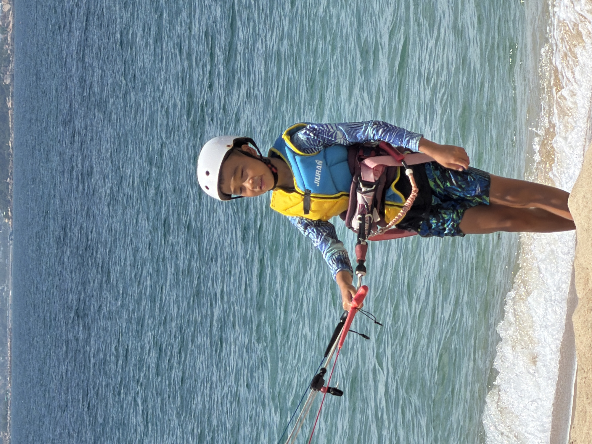 A young boy, wearing a white helmet, a yellow and blue life vest over a blue long-sleeved rashguard, and patterned shorts, stands on the sandy shore of the sea. He is holding a kiteboarding control bar. The water is choppy and blue, with a hint of green. In the background, a distant, hilly coastline is visible under a clear sky. The scene suggests a recreational activity, likely kiteboarding, taking place on a sunny day at a beach in Nha Trang, Vietnam.