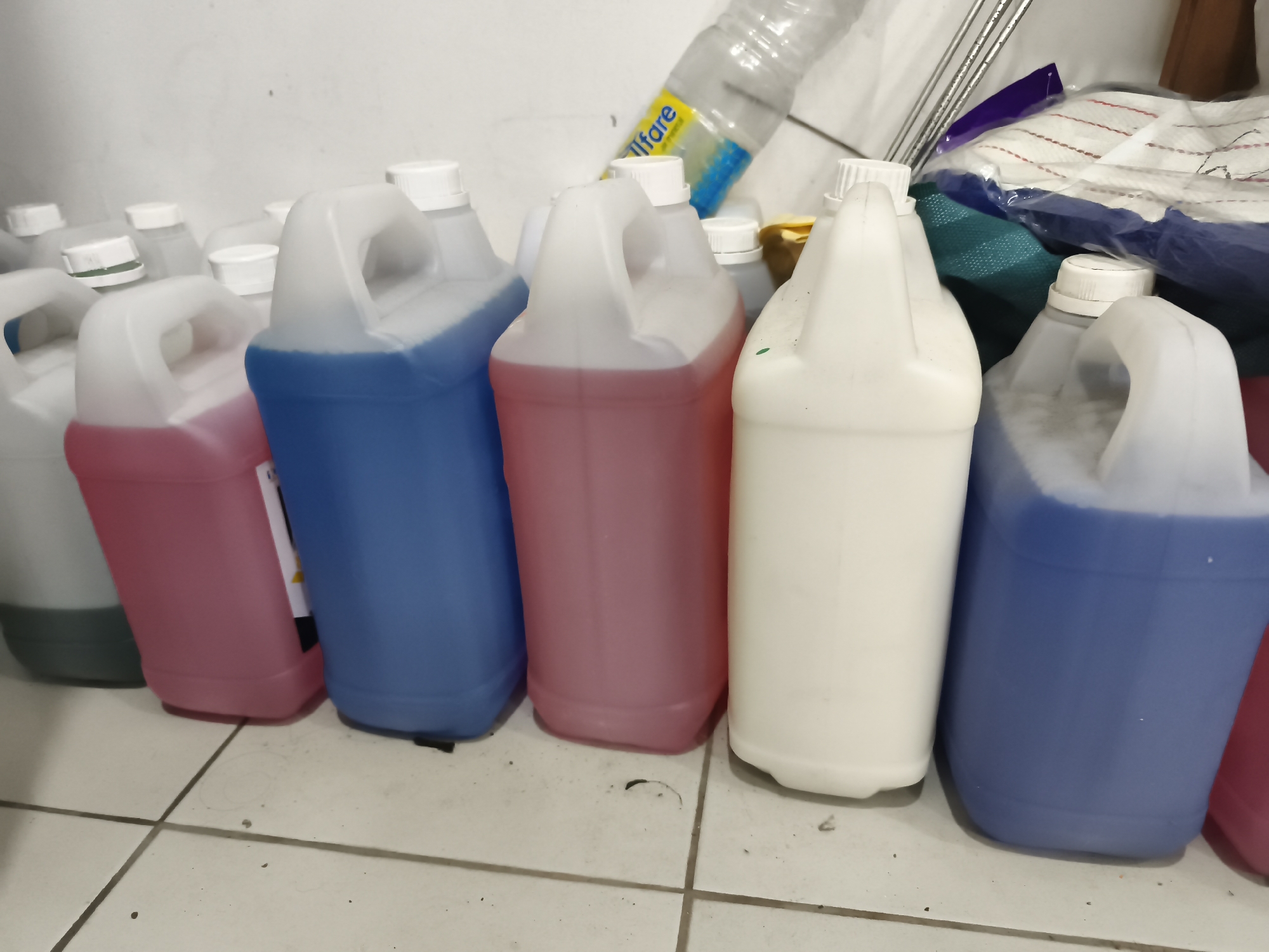 In Semarang, Indonesia, a collection of large plastic jugs filled with various colored liquids sits on a tiled floor against a white wall. The jugs are of different colors, including pink, blue, and white, and some are partially obscured by other items. A partially filled plastic bottle with a yellow label that reads "Ilfare" is visible in the background, leaning against the jugs. Other objects, such as fabric and metal rods, are also present in the background, suggesting a storage or workshop area. The lighting is dim, and the overall scene is cluttered.