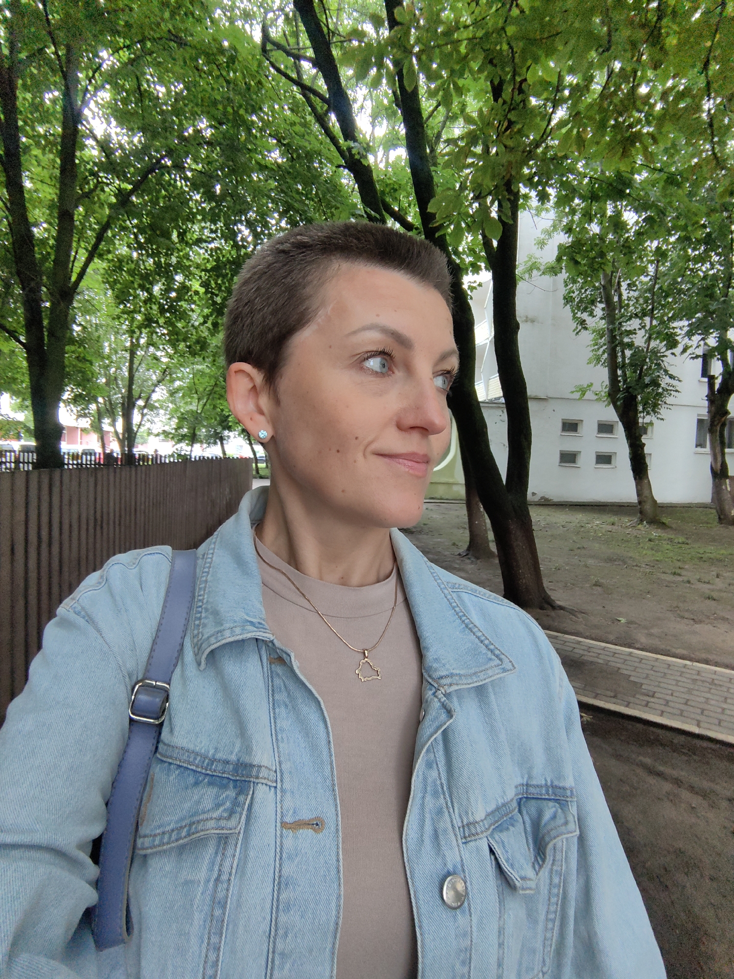 A woman with short, dark hair and blue eyes is captured in a close-up, selfie-style photo. She is wearing a light blue denim jacket over a tan shirt and a delicate gold necklace with a geometric pendant. A light blue strap from a bag crosses her chest. The background is an outdoor urban setting with mature trees displaying lush green foliage, a wooden fence, and a white building with several windows. A paved walkway is visible in the lower right portion of the image. The lighting suggests an overcast day. The scene is likely a park or residential area within the city of Minsk, Belarus.