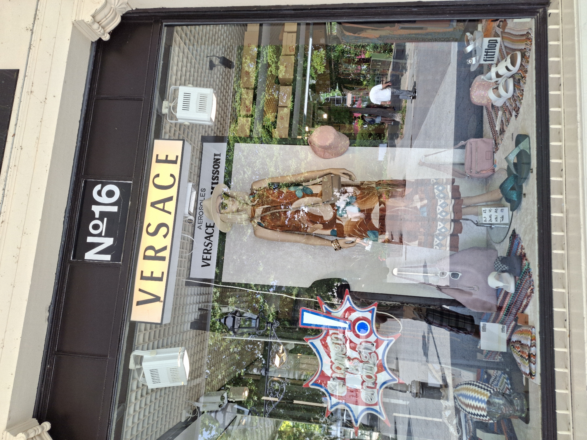 The image shows a retail store window display featuring mannequins and merchandise. The store is identified by signage as "N°16" and "VERSACE". A prominent sign in the window reads "Chosen is enough!". On display are dresses, hats, handbags, and footwear. The merchandise is arranged on shelves and the floor of the display. In the background, through the window, a street scene is visible with pedestrians and trees. The store is located in the city of Baden-Baden, Germany.