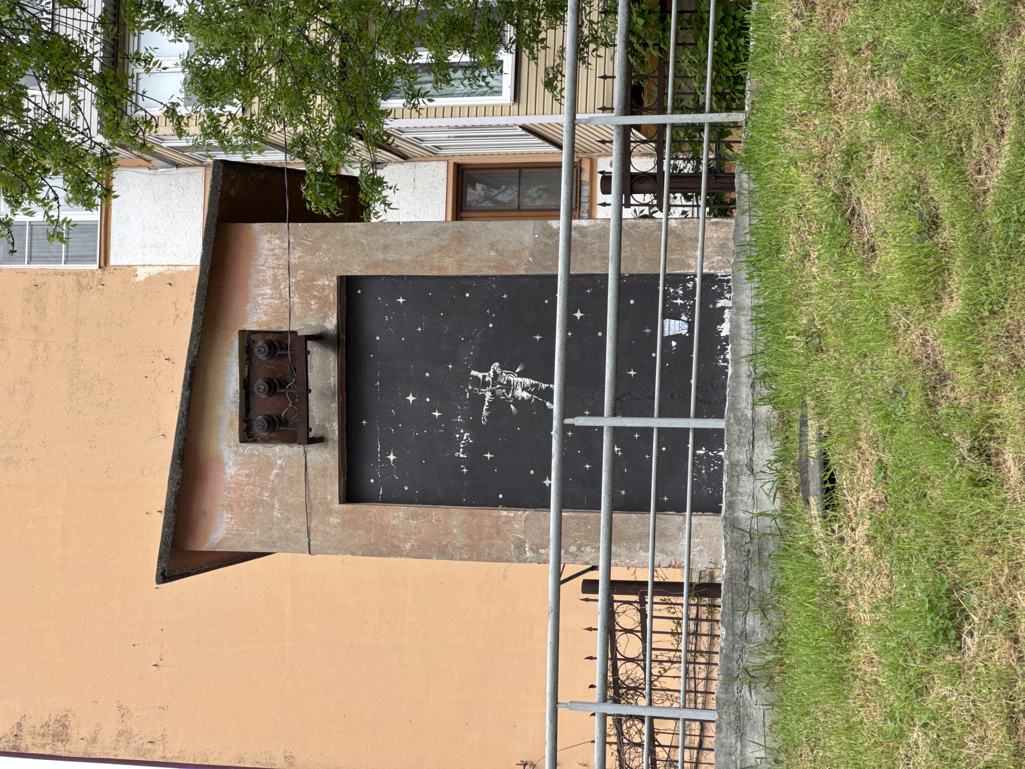 The image shows a section of a building with an adjacent grassy area. A metal fence partially obscures the view. On the building, there's an abstract mural painted on a recessed black rectangular area. The mural appears to depict an astronaut or figure in space amongst stars. Above the mural, there's a rusty electrical box with exposed wires. The building is painted in shades of orange and tan. The upper floors of the building, visible above the mural, show windows and different architectural styles. To the right of the building is a patch of green grass. The image provides no information about Zamankul, Russia, or any people, actions, or interactions at the location.