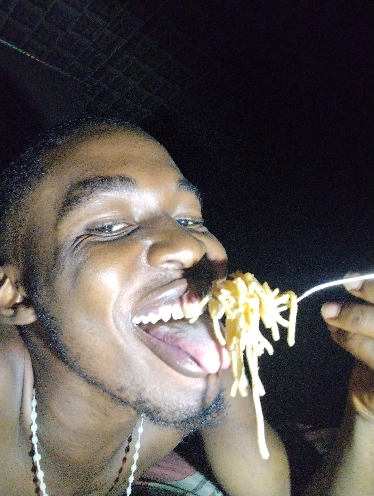 A close-up, low-light photograph features a Black man joyfully eating pasta. He is positioned in the foreground, with his face well-lit against a dark background, which exhibits a subtle grid-like pattern in the upper left corner. The man is looking directly at the camera with a wide, open-mouthed smile, revealing his teeth and tongue, and his eyes are crinkled at the corners, conveying an emotion of happiness or amusement.
He has short, dark, curly hair and stubble on his chin and jawline. Around his neck, a white beaded necklace or chain is visible. He holds a fork with his right hand, from which a generous portion of light-colored pasta, resembling spaghetti or noodles, is suspended, poised to enter his open mouth. The strong, direct lighting illuminates his features and the sheen on his skin, contrasting with the dark surroundings. The overall impression is one of candid enjoyment. The dark background suggests the photo was taken at night or in a dimly lit indoor setting. There are no visible location cues within the image. No text is visible.