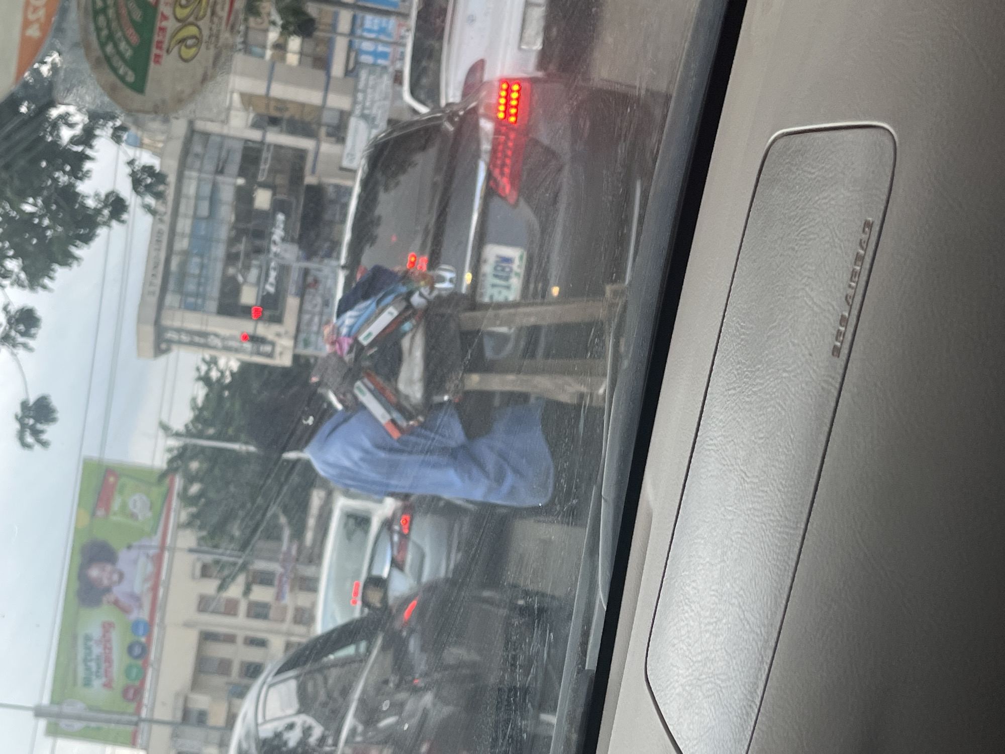 In Abuja, Nigeria, a street vendor stands beside a traffic jam. They wear a blue shirt and dark trousers, holding an assortment of items. The vendor is positioned near a dark-colored Honda sedan with license plate G-14BW. Behind the Honda, a white car is visible. In the background, a billboard displays an advertisement with a child. The scene unfolds in an urban environment with visible traffic lights and buildings.