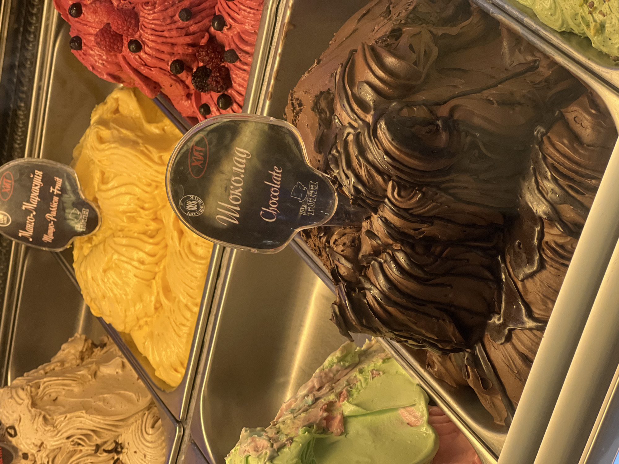 A tilted high-angle close-up shot shows a display case containing several flavors of ice cream. The ice cream flavors visible are red with berries, yellow, brown, green with pink accents, and a light brown swirl. Labels indicate the flavors: "Mango-Passion Fruit" (possibly in Russian, "Манго-Маракуйя") and "Chocolate" (also possibly in Russian, "Шоколад"). The labels feature the brand name "Mr. TUTTI." The presence of Russian text suggests that this is likely in Moscow, Russia. The overall impression is a colorful display of artisan gelato.