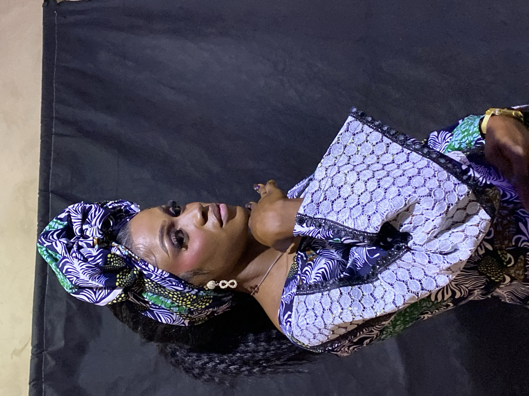 This portrait, taken in Kuru Dungus, Nigeria, features a woman posing against a dark fabric backdrop, with a lighter wall visible to the left. The woman, with dark skin, is depicted from the chest up, looking slightly upwards and to her right with a serene and poised expression.
She is dressed in a vibrant, multi-patterned ensemble and a matching headwrap (gele). The fabric of her attire displays intricate designs in various shades of blue, white, green, and yellow. Her top features a section with a lace-like texture characterized by circular patterns. Her hair is styled in dark braids or twists, visible falling over her left shoulder.
For accessories, she wears prominent earrings resembling an infinity symbol, adorned with sparkling stones. A delicate silver-colored chain necklace is visible around her neck. A gold-colored watch is on her right wrist, and her fingernails are painted a dark hue. Her makeup includes defined eyebrows, dark eyeliner, and a soft lip color, enhancing her features. Her right hand is gently raised, resting near her chest or shoulder. The lighting suggests a controlled indoor environment, highlighting her features and the textures of her attire. No visible text is present in the image.