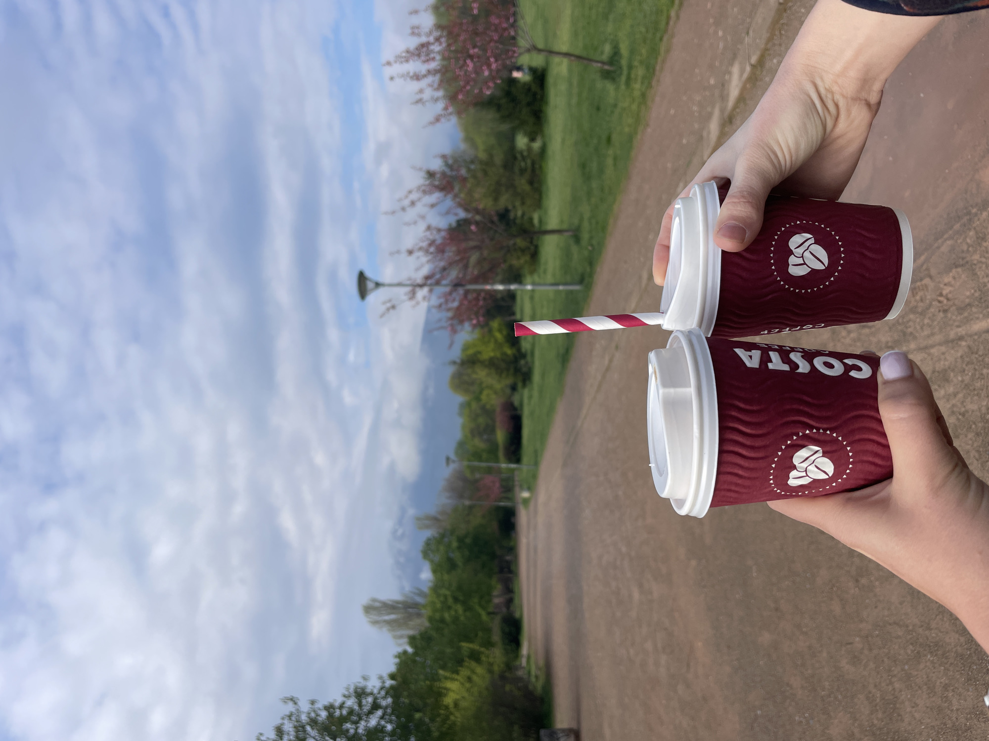 A person holds two Costa Coffee cups with red and white straws in a park setting in Sofia, Bulgaria. The cups are the focal point, with the Costa Coffee logo clearly visible. The background features a park landscape with trees, grass, and a pathway. The sky is partly cloudy. No specific landmarks are visible to definitively pinpoint the exact location within Sofia.