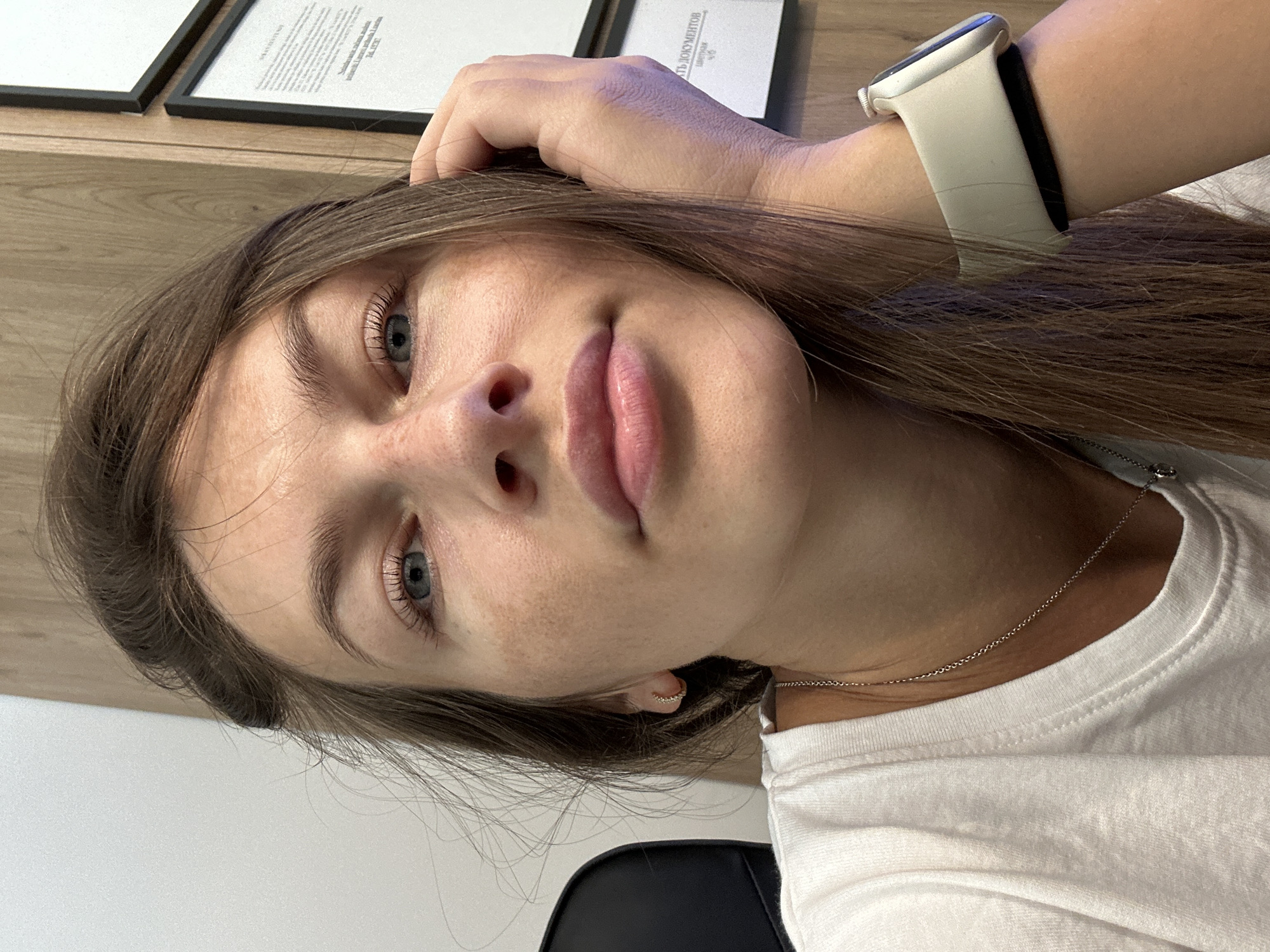 A young woman is captured in a close-up, portrait-style photograph, her head tilted and her hand resting on her forehead. She has long, brown hair that cascades around her face and shoulders. Her eyes are a striking blue, and her lips are slightly parted, conveying a thoughtful or perhaps playful expression. She is wearing a white t-shirt and a delicate silver necklace. On her left wrist, she sports a white Apple Watch.
The background suggests an indoor setting. To the upper left of the frame, two framed documents are visible on a wooden surface, possibly a desk or shelf. One document appears to have Cyrillic text. The overall lighting is natural and soft, hinting at daytime.
The image is oriented sideways, which may be a stylistic choice or accidental. The framing is tight on the woman's face and upper body, creating an intimate feel. The scene appears to be in a personal or professional space, potentially an office or home. There are no overt indications of the weather or a specific time of day beyond it being daylight. The woman's expression is neutral to slightly curious.