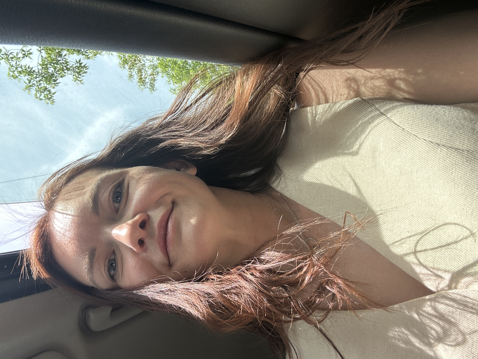The image shows a woman with light skin, reddish-brown hair, and light-colored eyes. She is in a car, looking towards the camera with a slight smile. The window is visible in the upper left corner, showing trees and a cloudy sky. The interior of the car is dark, with a light-colored seat visible. There is no information available to determine the location as the city of Chaikovsky, Russia.