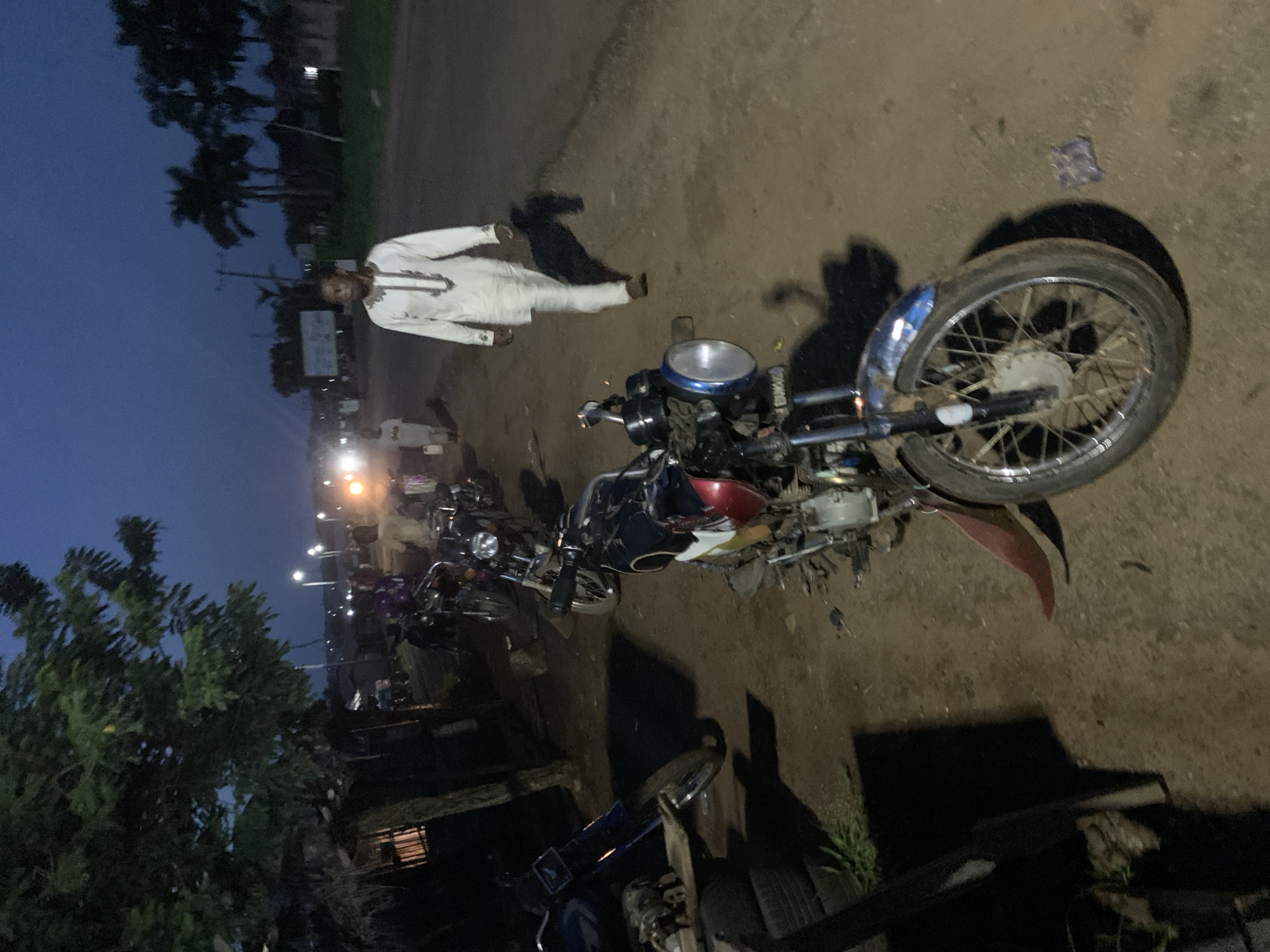 The image depicts a scene in Gbeleko, Nigeria, at dusk. A man wearing a white traditional garment stands on a dirt road adjacent to several parked motorcycles. A motorcycle in the foreground is the most prominent feature, showing wear and possible disrepair. Streetlights illuminate the background, where other motorcycles are parked. The environment includes trees and vegetation characteristic of the region. The sky is a dark blue, suggesting the early evening.