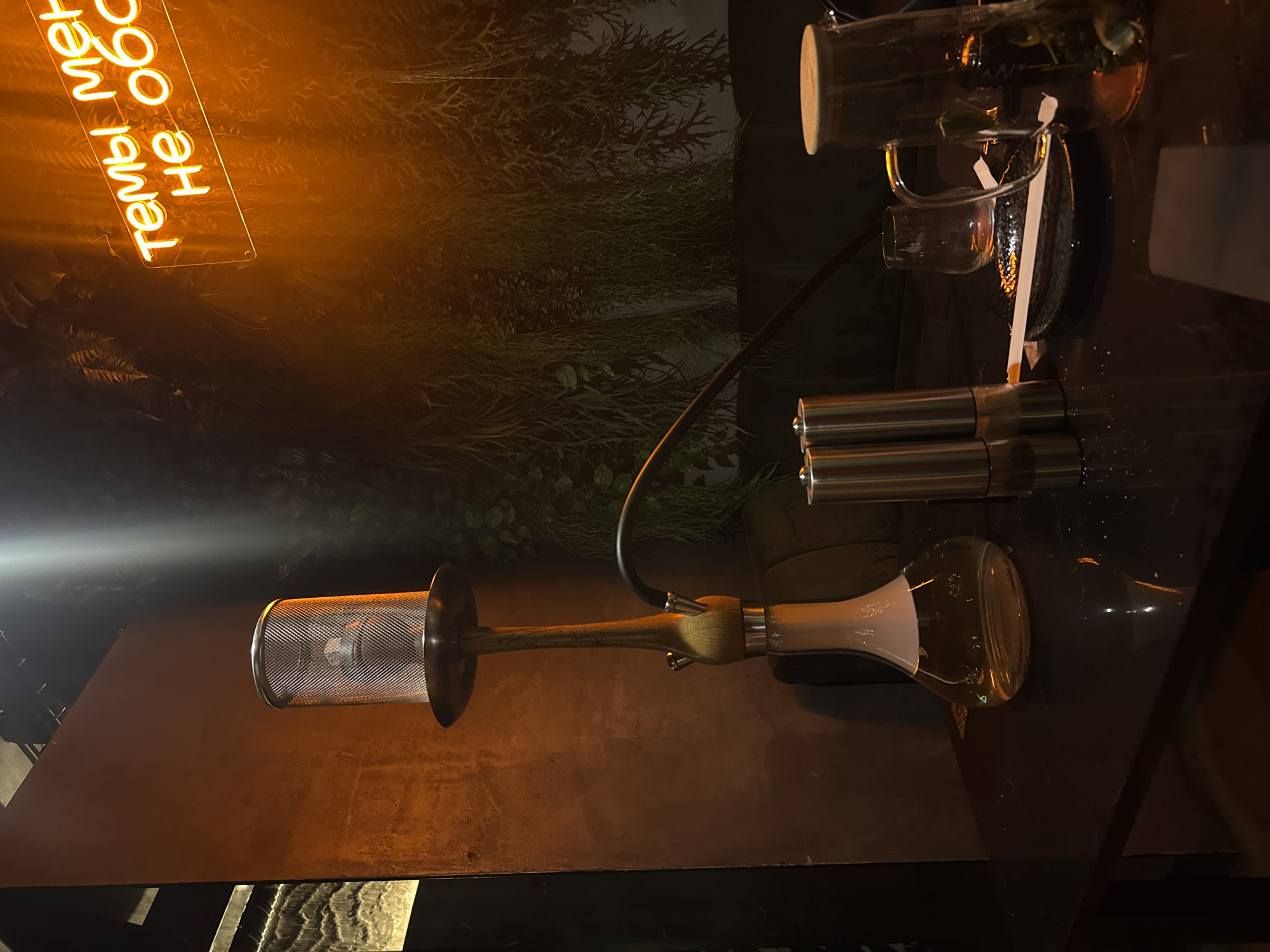 The image shows a hookah on a table, with a neon sign in the background. The hookah has a glass base and a metal stem, with a mesh bowl at the top. The neon sign is orange and says "Тем, что меня не обо". The background is dark, with some green plants visible. The time of day is likely evening or night, as the neon sign is illuminated. The setting appears to be indoors, possibly a bar or lounge. There are no people visible in the image. The overall mood is dimly lit and somewhat mysterious. The text is in Russian.