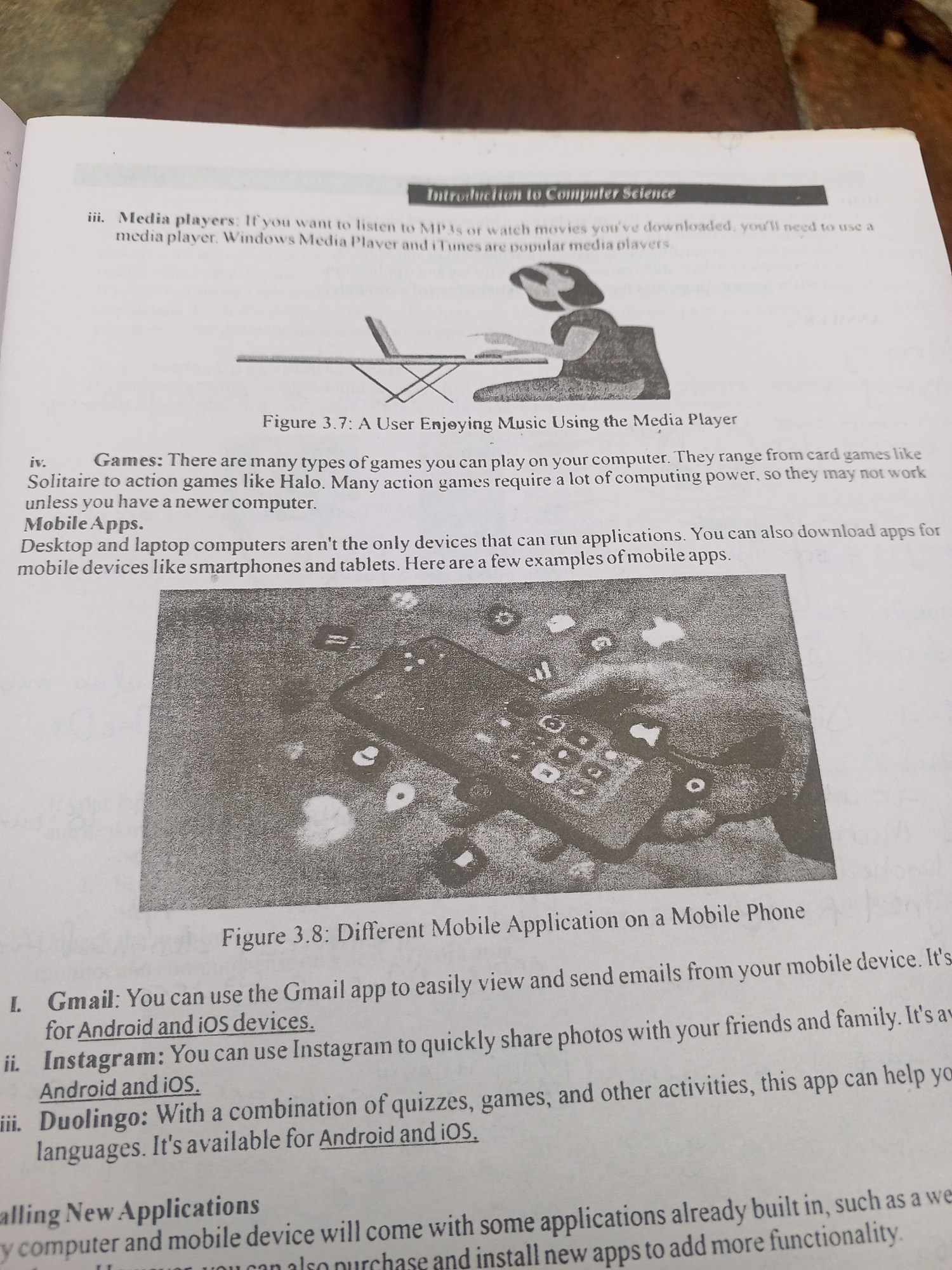 This media file is a close-up, angled shot of an open book. The book is about computer science, specifically detailing different applications on mobile phones and computers. The page features text and images that are black and white. The text discusses media players, games, mobile apps, and specific applications like Gmail, Instagram, and Duolingo, along with their availability on Android and iOS devices. It also mentions installing new applications. There are two figures depicted in the book: Figure 3.7 shows a person using a media player, and Figure 3.8 displays a hand interacting with a smartphone screen, which is covered in app icons. The background of the image is out of focus, showing the textures of what appears to be fabric, possibly clothing or a textile covering. The lighting suggests an indoor setting, and the overall tone of the image is educational. No specific time of day, weather, or location cues from Jalingo, Nigeria are visible within the frame of the book itself.