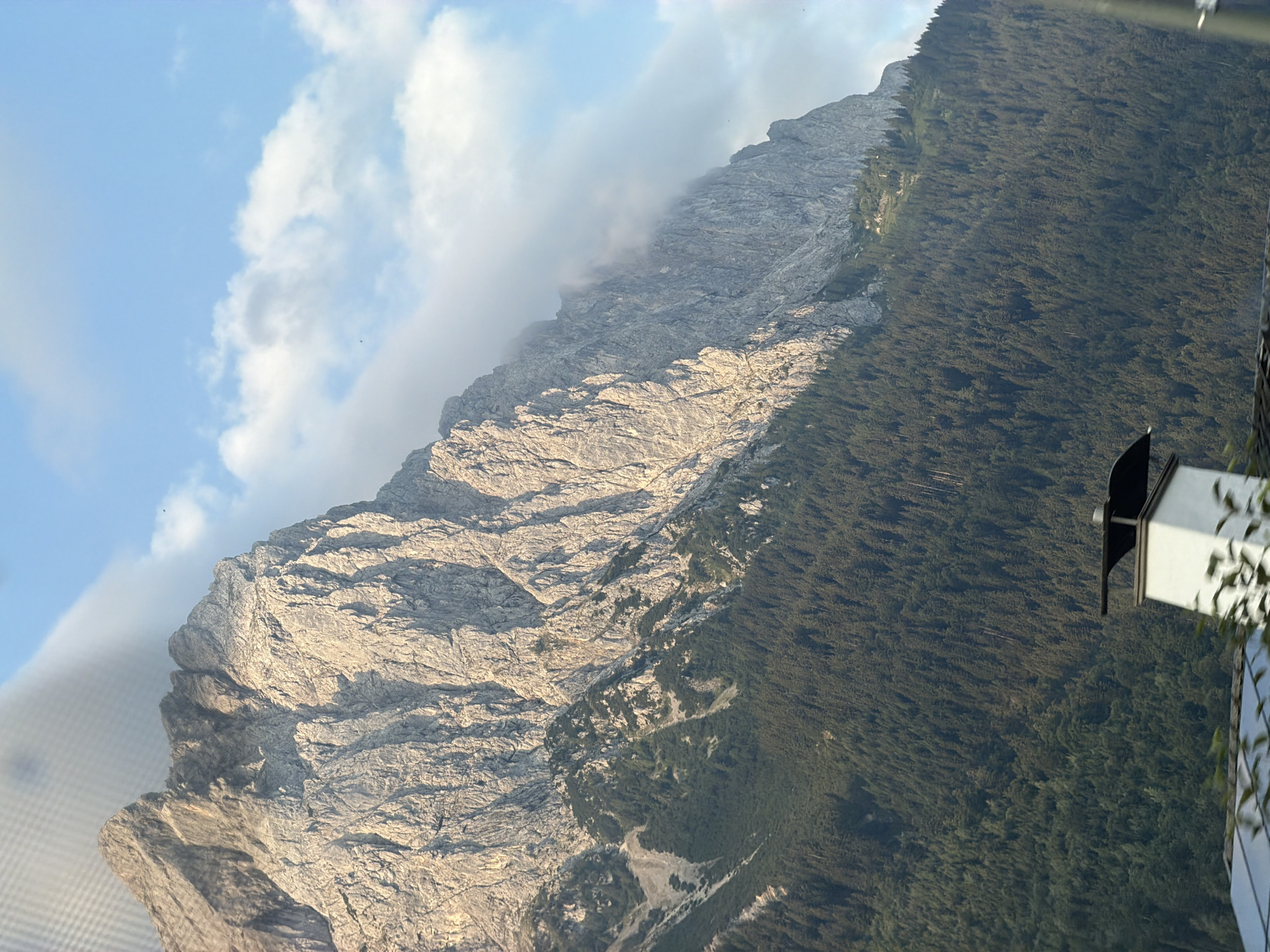 A bright, sunlit mountain range dominates the frame. The rugged, grey rock face of the mountain is textured with deep crevices and sharp ridges, casting shadows that emphasize its formidable stature. Patches of white clouds cling to the upper slopes, partially obscuring the peak and lending a dramatic, ethereal quality to the scene. Below the rocky summit, a dense, dark green forest covers the lower mountain slopes, creating a stark contrast with the lighter rock above. The trees are closely packed, forming an unbroken canopy that stretches across the hillside. In the foreground, a portion of a white building with a black-capped chimney is visible, with a few out-of-focus green leaves obscuring the bottom edge of the image. The sky above is a clear, pale blue, with a few scattered white clouds. A faint grid pattern is visible in the upper left corner, suggesting the image was taken through a window. The scene depicts a natural mountainous landscape.