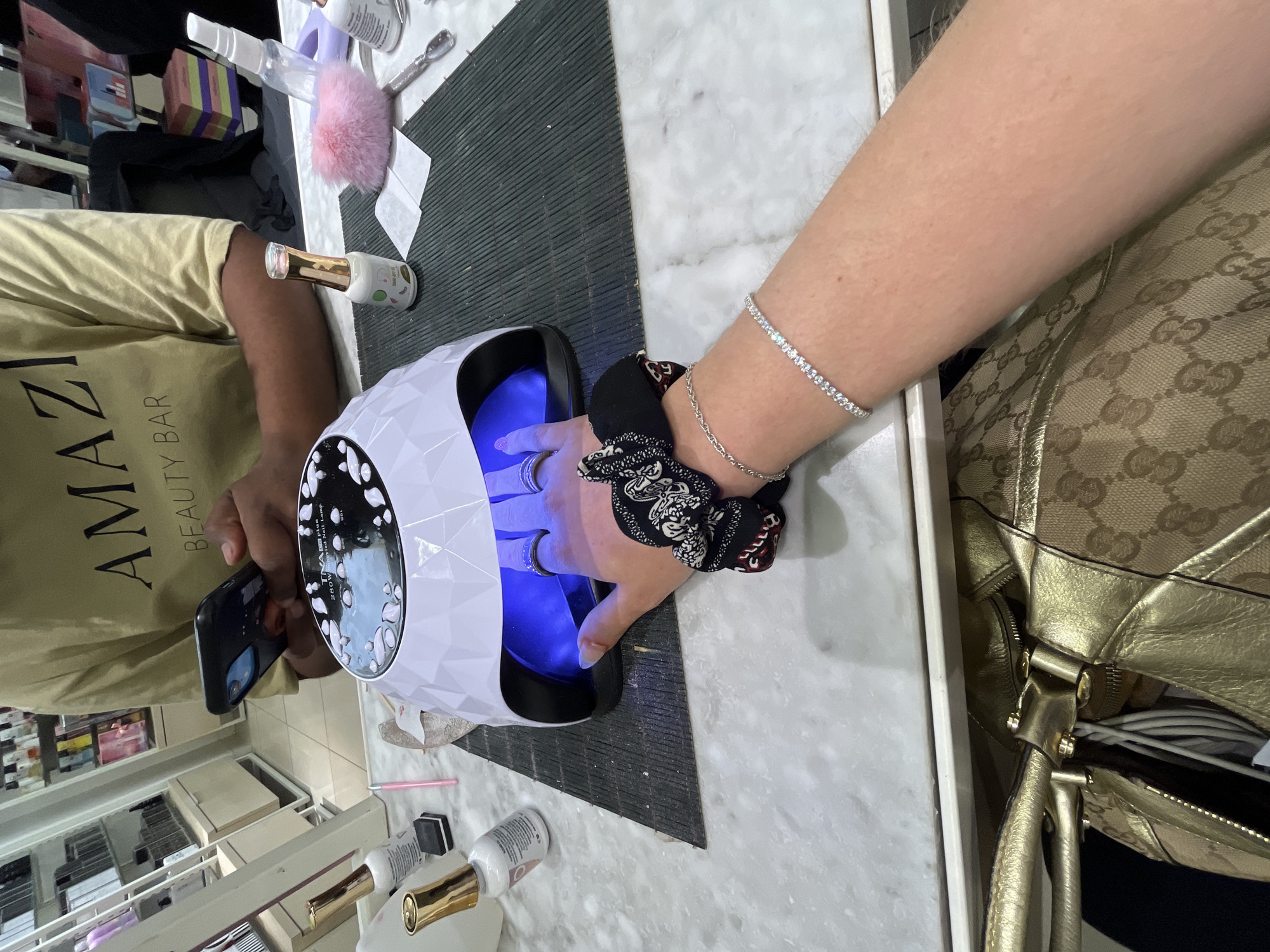 A person is having their nails done, placing their hand in a UV nail lamp for gel curing. A manicurist, wearing a tan t-shirt with "AMAZI BEAUTY BAR" printed on it, is operating the lamp and possibly taking a picture with their smartphone. The person getting their nails done is wearing a black and white patterned scrunchie and a tennis bracelet. A handbag is resting nearby. The setting appears to be a nail salon, based on the bottles of nail polish, tools, and shelving of products visible in the background. This is likely at Amazi Beauty Bar in Johannesburg, South Africa.