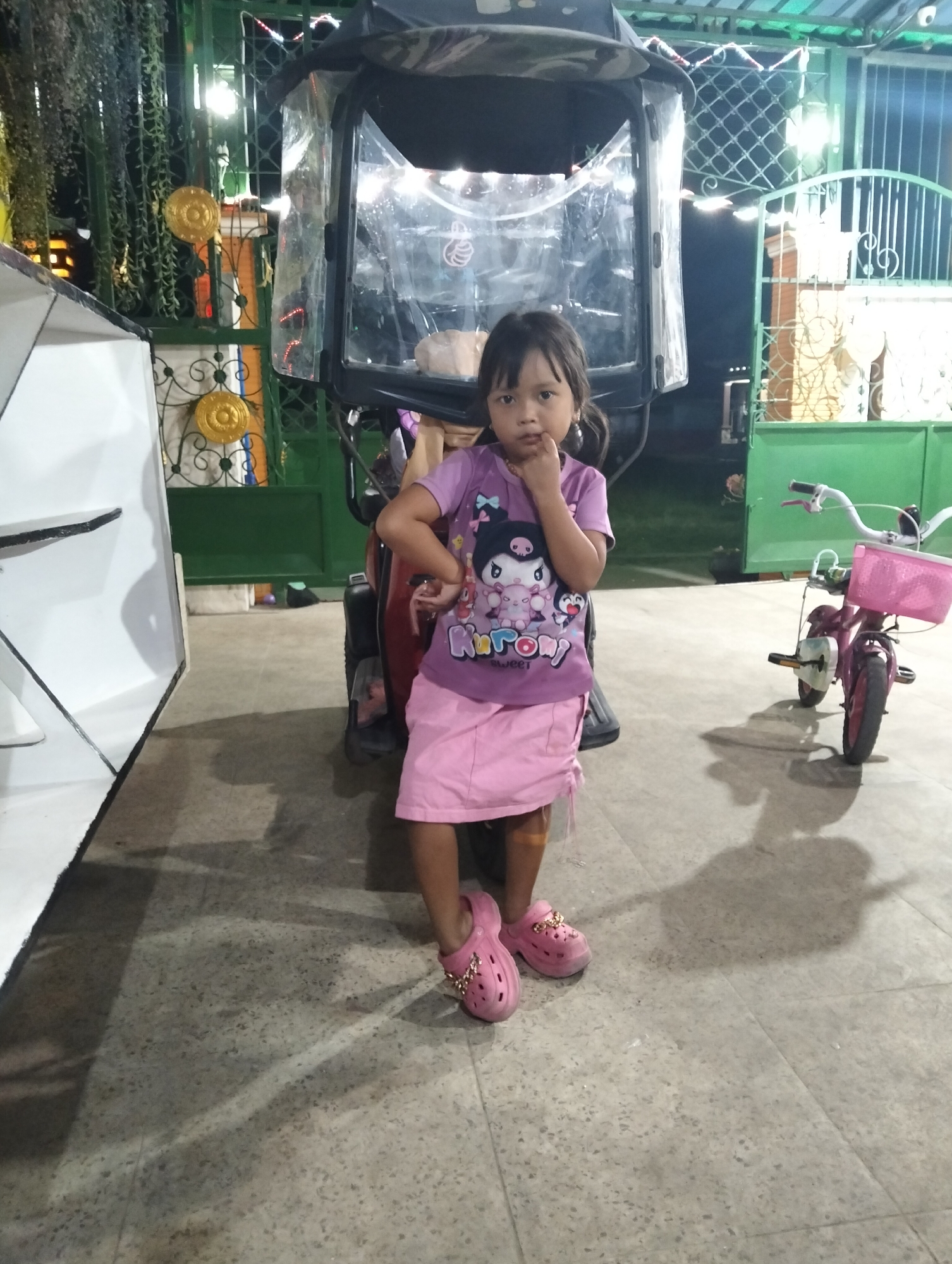 A young girl stands in front of a vehicle, possibly a motor tricycle, with a pink bicycle to her right. She is wearing a purple t-shirt with a cartoon character and a pink skirt. Her feet are clad in pink crocs. The scene appears to be outdoors at night, with artificial lighting illuminating the area. In the background, a green metal fence and some hanging plants are visible. The ground is paved with textured tiles. This image was taken in the city of Muncar, Indonesia.