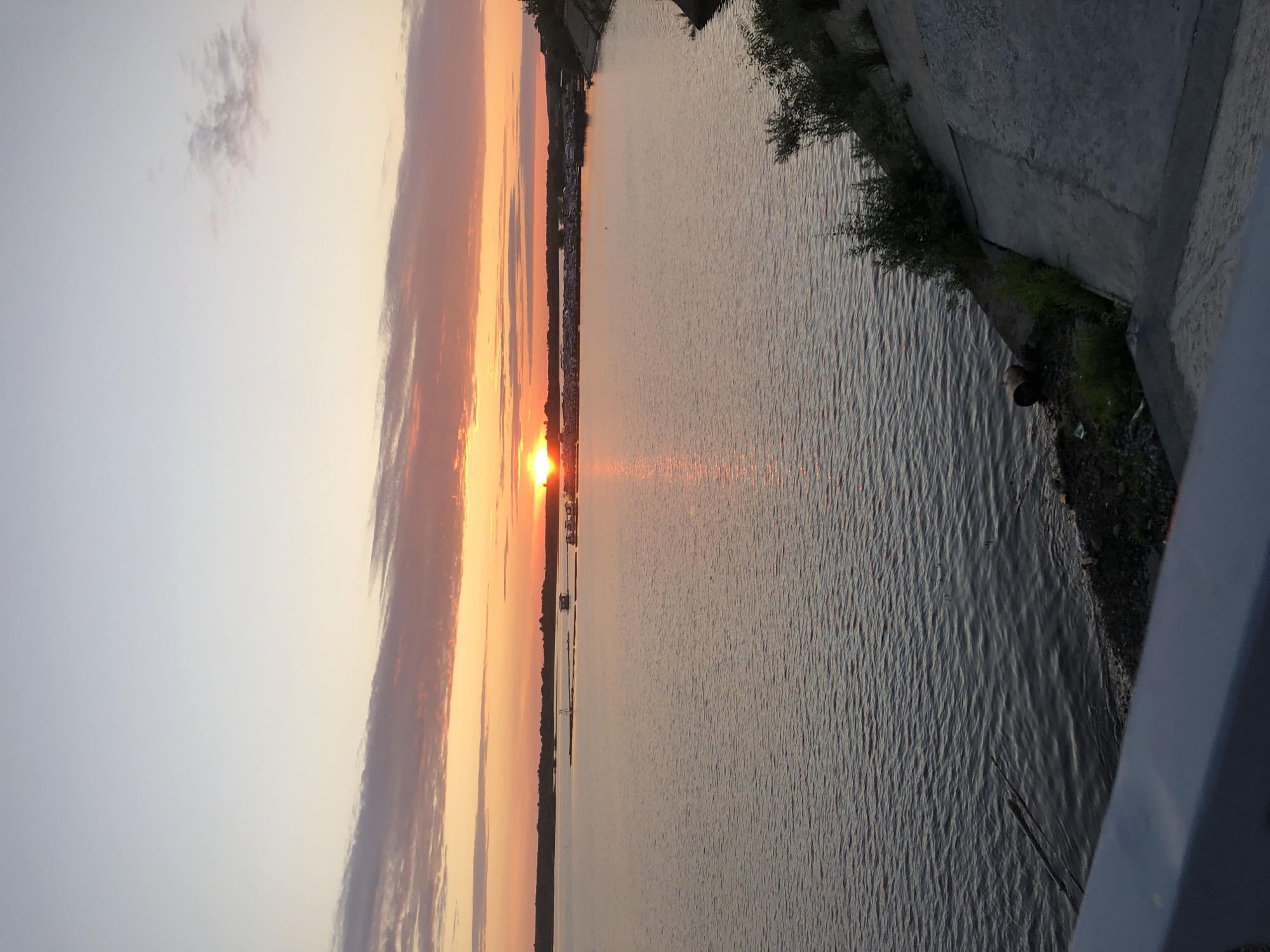 This is a vertical, landscape photograph of a body of water with a sunset in the background. The sun is low on the horizon, casting a warm orange and pink glow across the sky and reflecting on the water. Clouds are visible in the sky, painted with the colors of the sunset.
The scene appears to be a river or lake with a shoreline. The shoreline has some vegetation and a concrete embankment. In the distance, along the far shoreline, there are some buildings that suggest a town or city. The water's surface is rippled, indicating a gentle breeze. The overall mood of the photograph is peaceful and serene, capturing the beauty of a sunset. There is no visible text. The time of day is sunset. The weather appears to be clear with some clouds. The location is Solikamsk, Russia.
