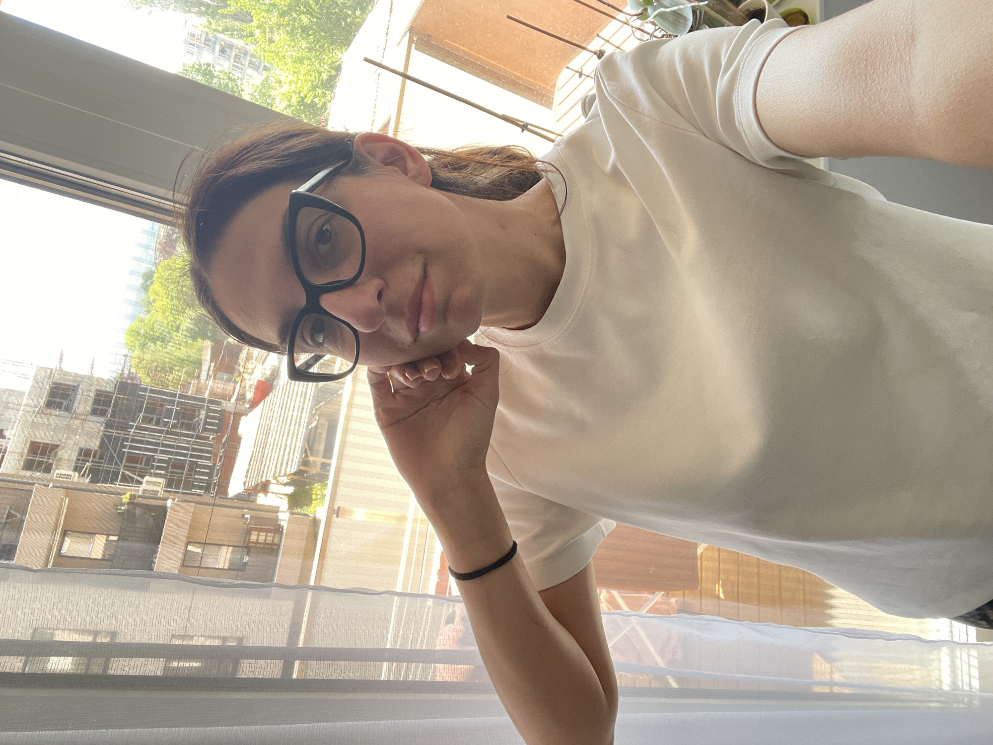 A young woman with dark hair and glasses poses for a selfie near a window. She wears a white t-shirt and has a black elastic band on her left wrist. Her head rests on her hand, and she looks towards the camera. Outside the window, buildings are visible, including one with scaffolding. This cityscape suggests the image was taken in an urban area. It is not possible to identify the location as the city of Sofia, Bulgaria based solely on this image.