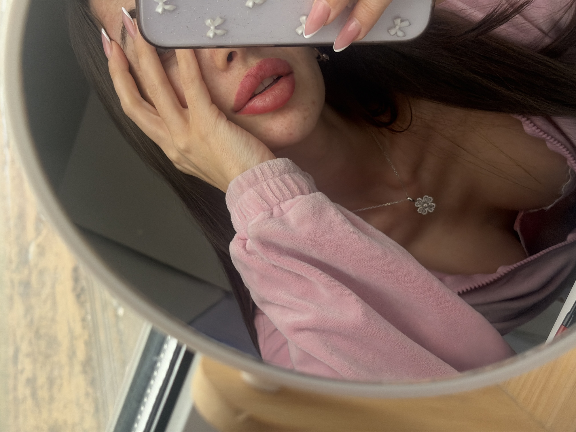 A female subject is depicted in a mirror selfie, partially obscuring her face with her left hand. Her long, dark hair is visible, falling over her right shoulder. She wears a pink, textured, zip-up top, exposing a silver chain with a floral or clover-shaped pendant on her chest. Her lips are prominent, enhanced with red lipstick, and the skin on her chin exhibits some texture. Both visible hands display manicured nails with a pink base and white French tips. The right hand holds a smartphone with a light-colored case decorated with small white bows, positioning it for the photograph. The mirror reflects a window in the upper left background, through which a blurred, light brown landscape with faint horizontal lines, indicative of railway tracks, is visible. The interior environment includes a light-colored, possibly wooden, surface at the bottom left, with a dark object resembling a pen or marker. The scene suggests the subject is aboard a train or in an area immediately adjacent to railway lines, within the broader vicinity of Saint Petersburg, Russia.
